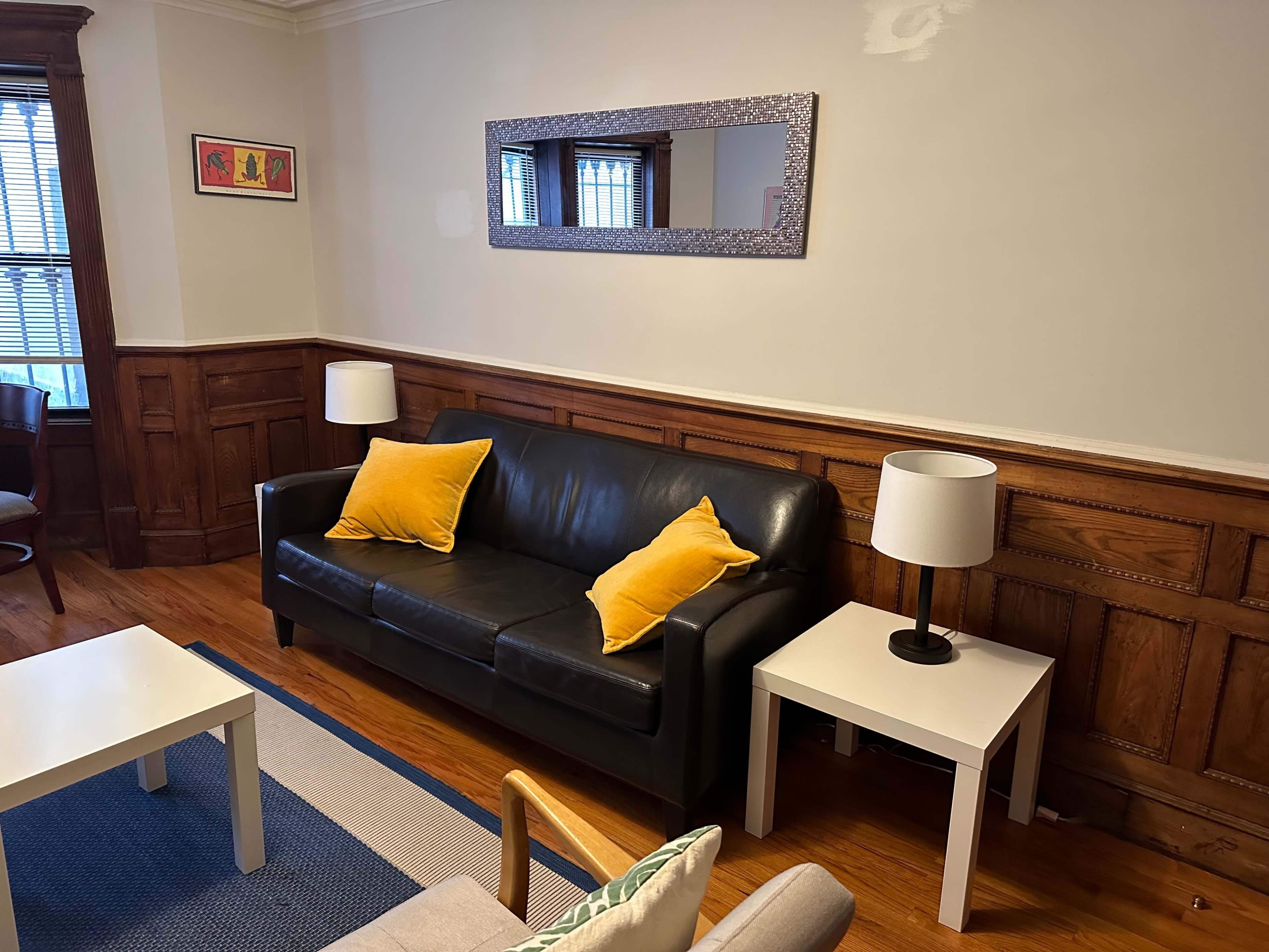 The living room features a black leather sofa with yellow pillows flanked by two white side tables with lamps, against a backdrop of wood-paneled walls.