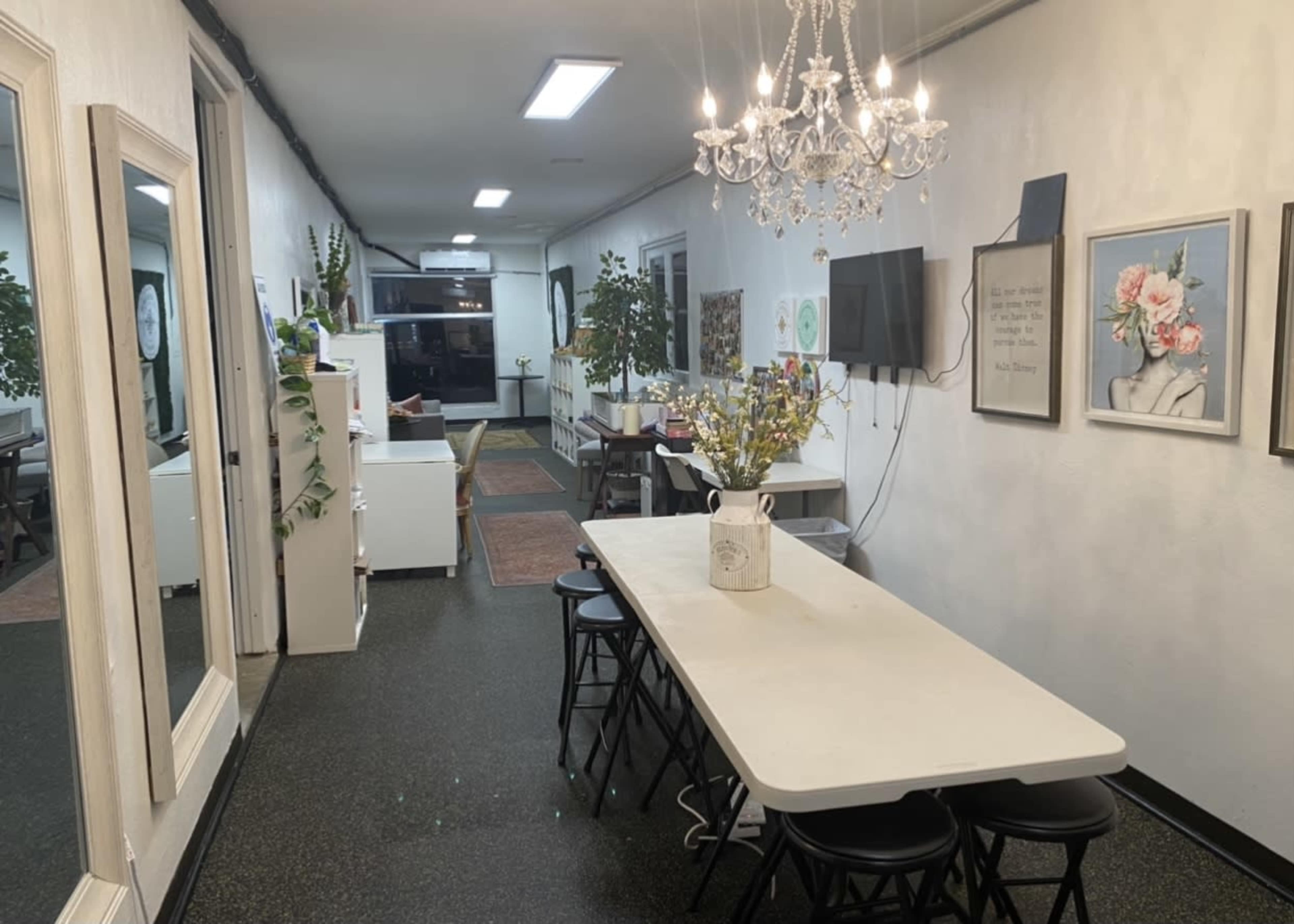 The image shows a well-lit hallway with a long white table surrounded by black stools, flanked by mirrors and decorative plants, leading to a living area with artwork on the walls.