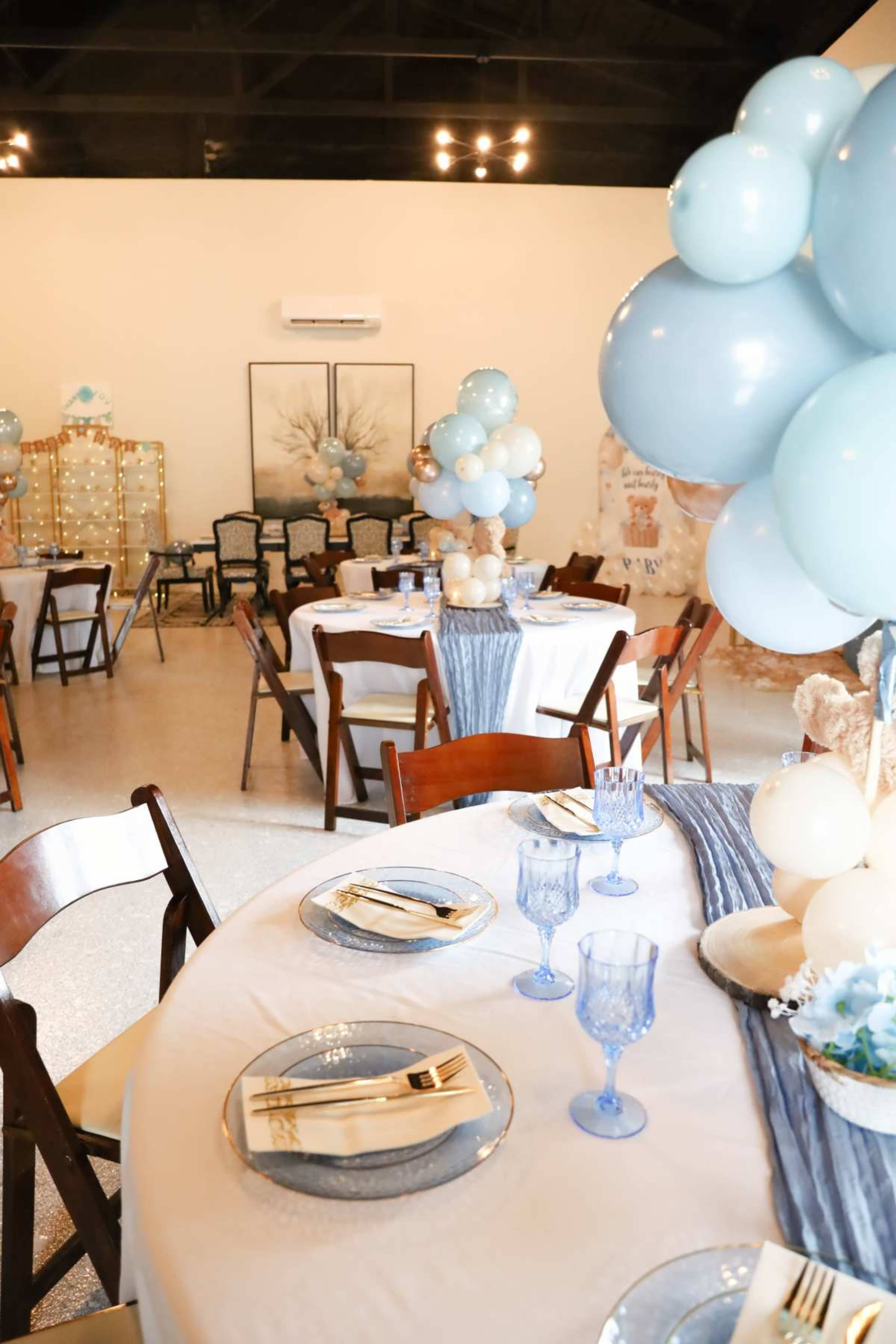 The image shows a decorated event space with tables set for a celebration, featuring blue and white balloons, elegant tableware, and chairs arranged around the tables.
