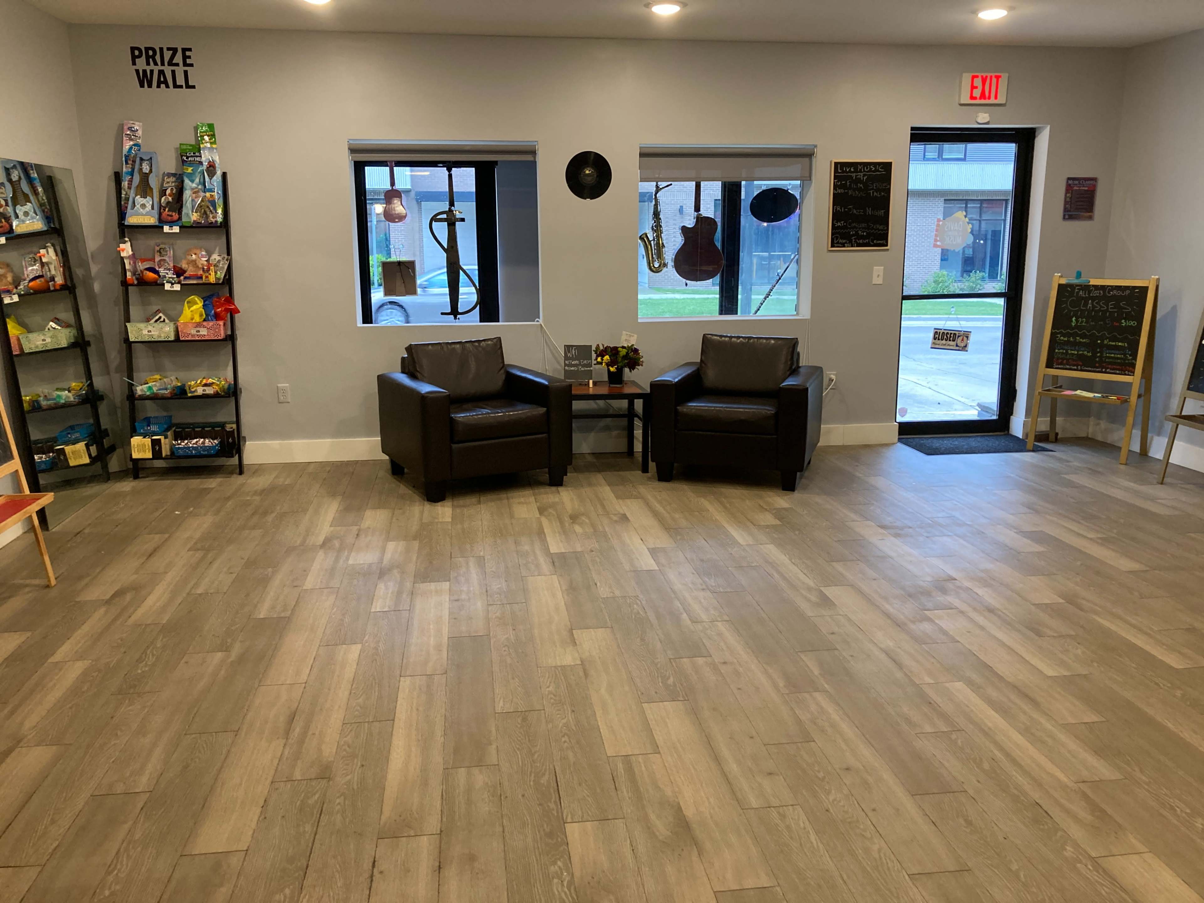 The image shows an inviting room with two dark chairs facing a small table, a prize wall displaying items on the left, and large windows letting in natural light.