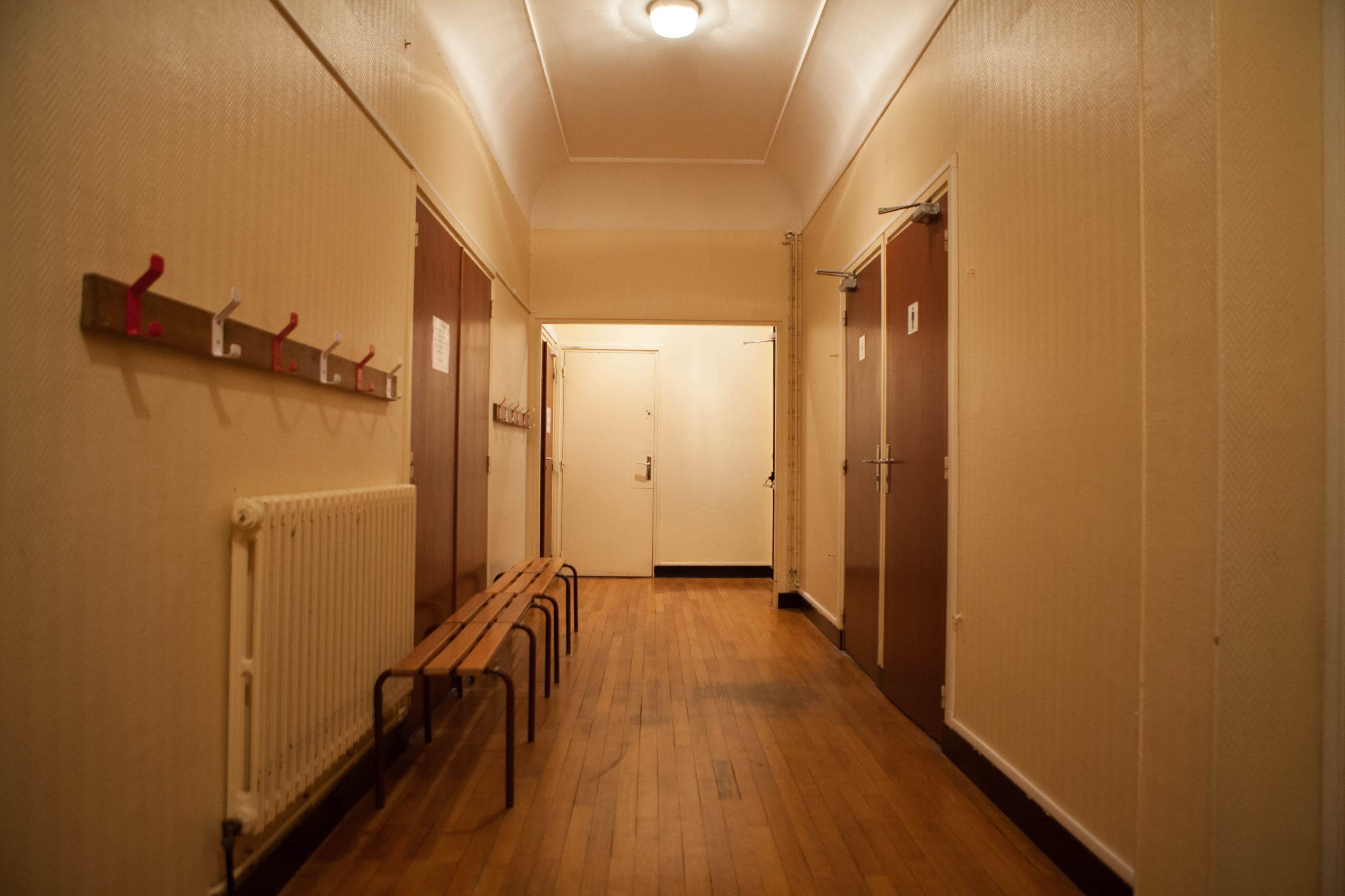The image shows a narrow hallway with wooden flooring, benches, and doors along the walls, along with coat hooks mounted on one side.