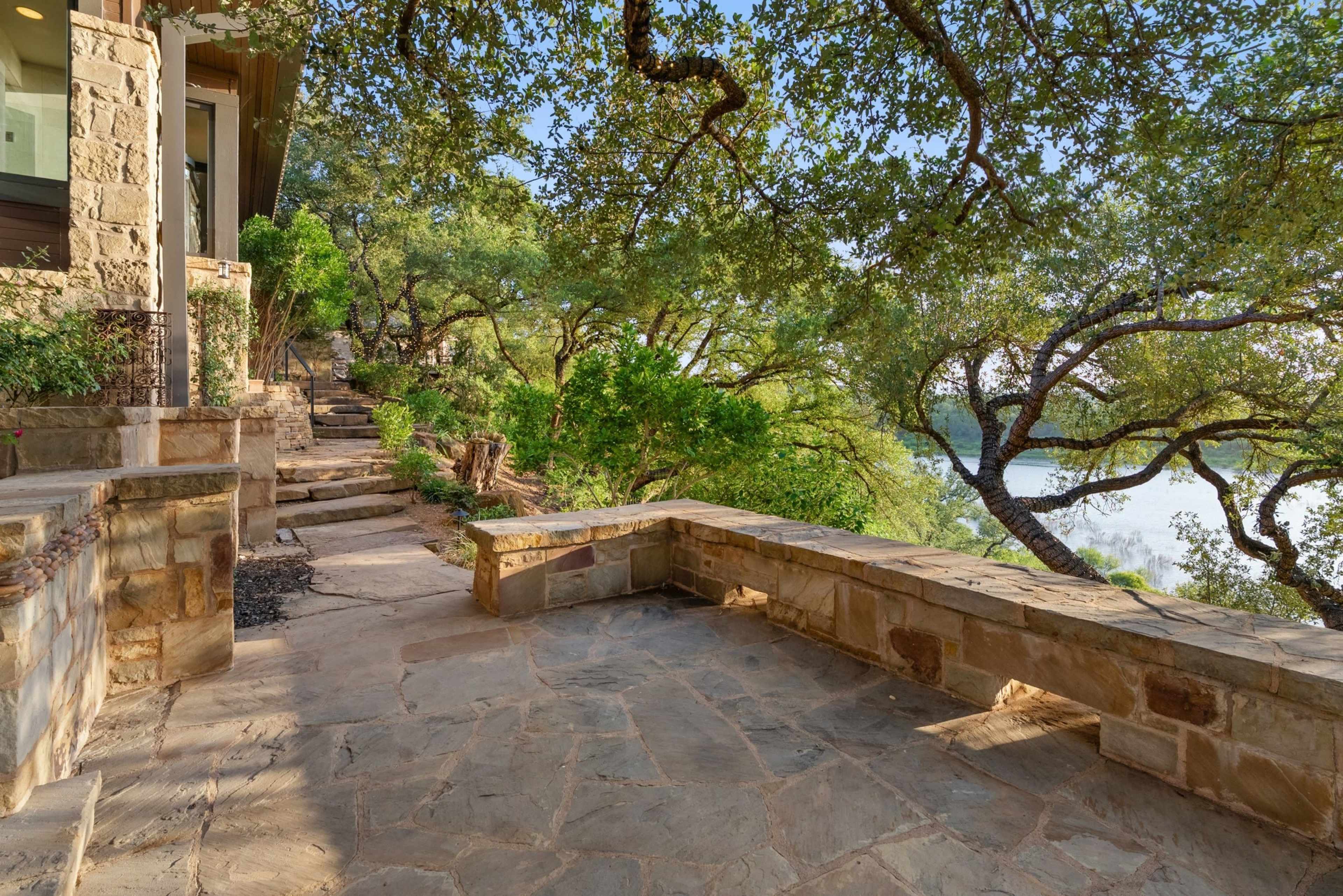 A stone pathway winds through an outdoor area surrounded by lush greenery and views of a lake.