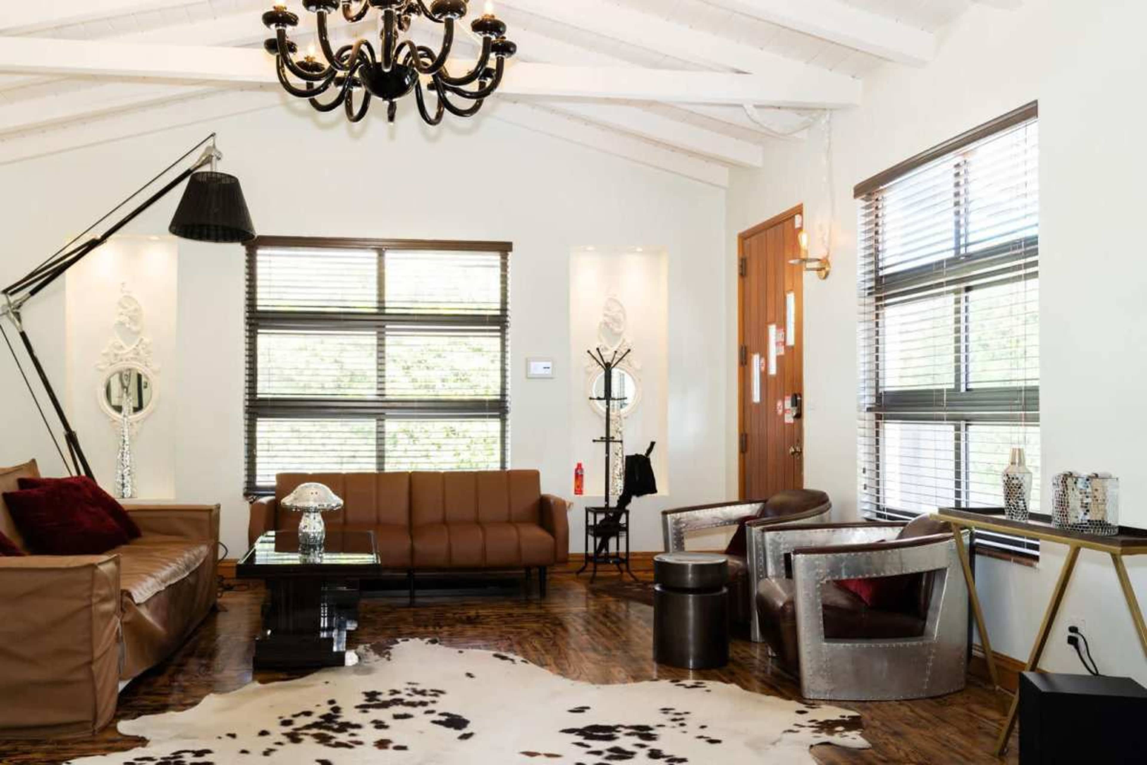 The living room features a plush brown sofa, two metal armchairs, a large chandelier, and a cowhide rug on a hardwood floor, with large windows allowing natural light.