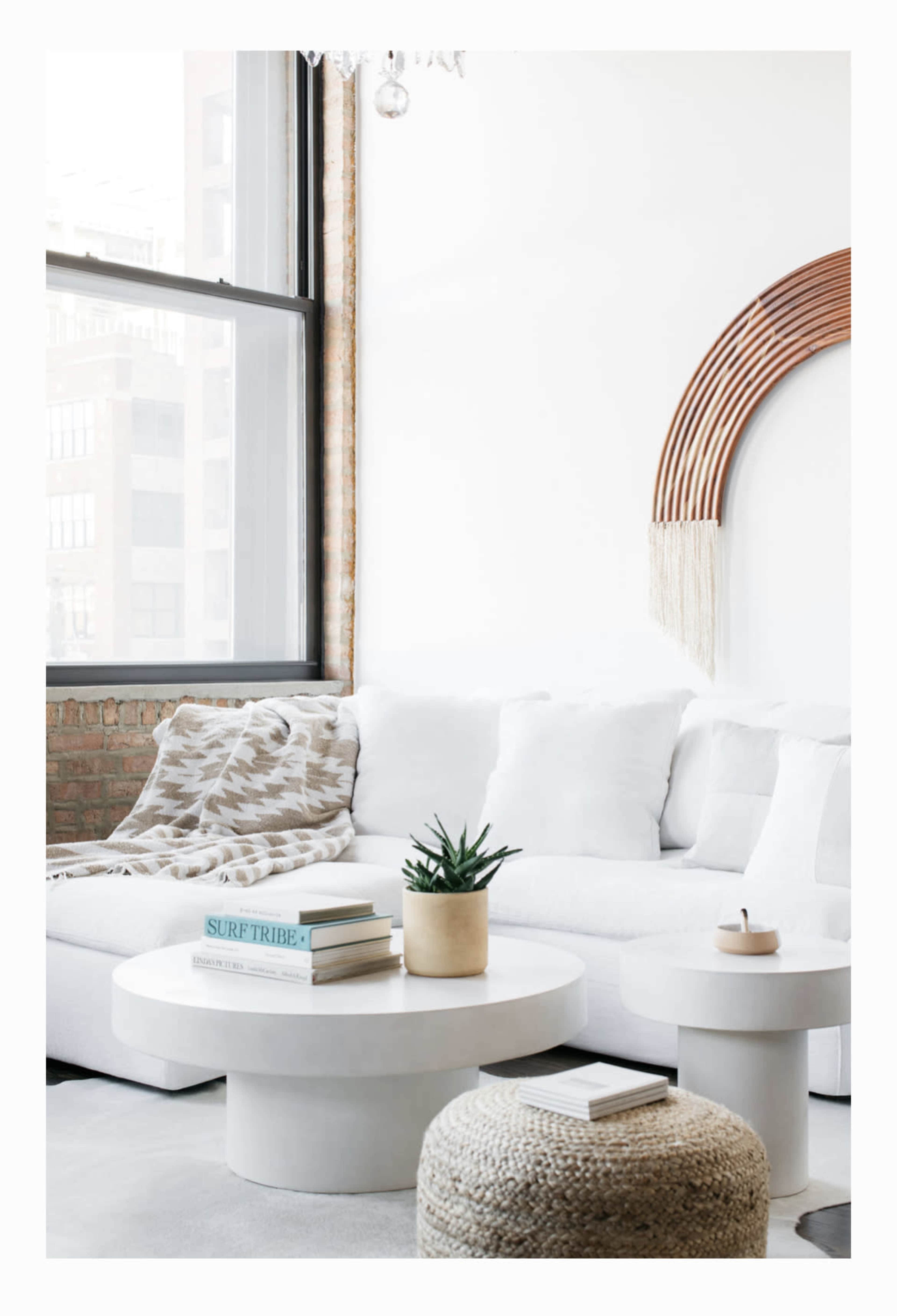A modern living room with a white sectional sofa, a round coffee table, a woven storage basket, and a decorative wall hanging, all complemented by large windows letting in natural light.