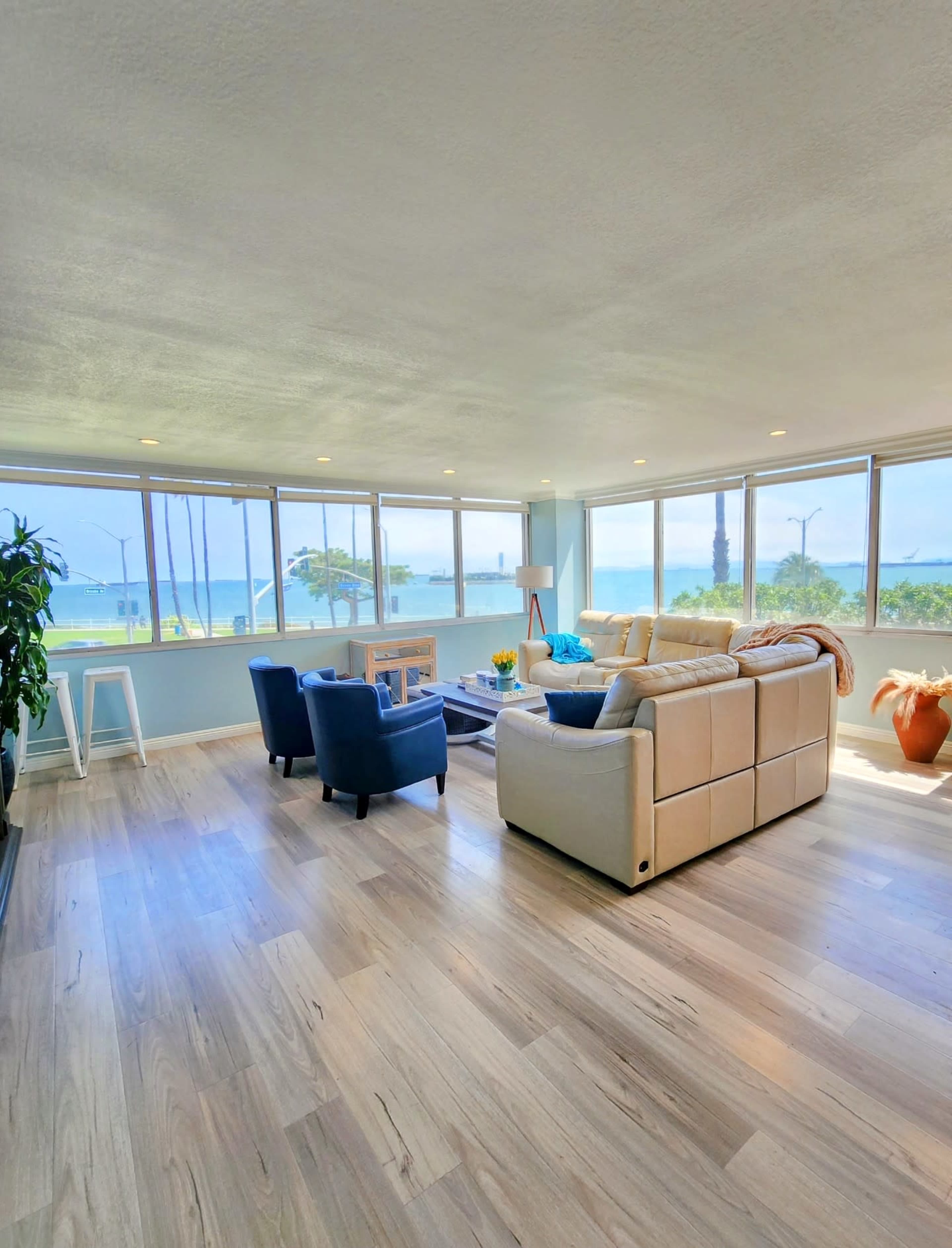 A living room features a white couch, blue chairs, and large windows that offer a view of the ocean and palm trees outside.