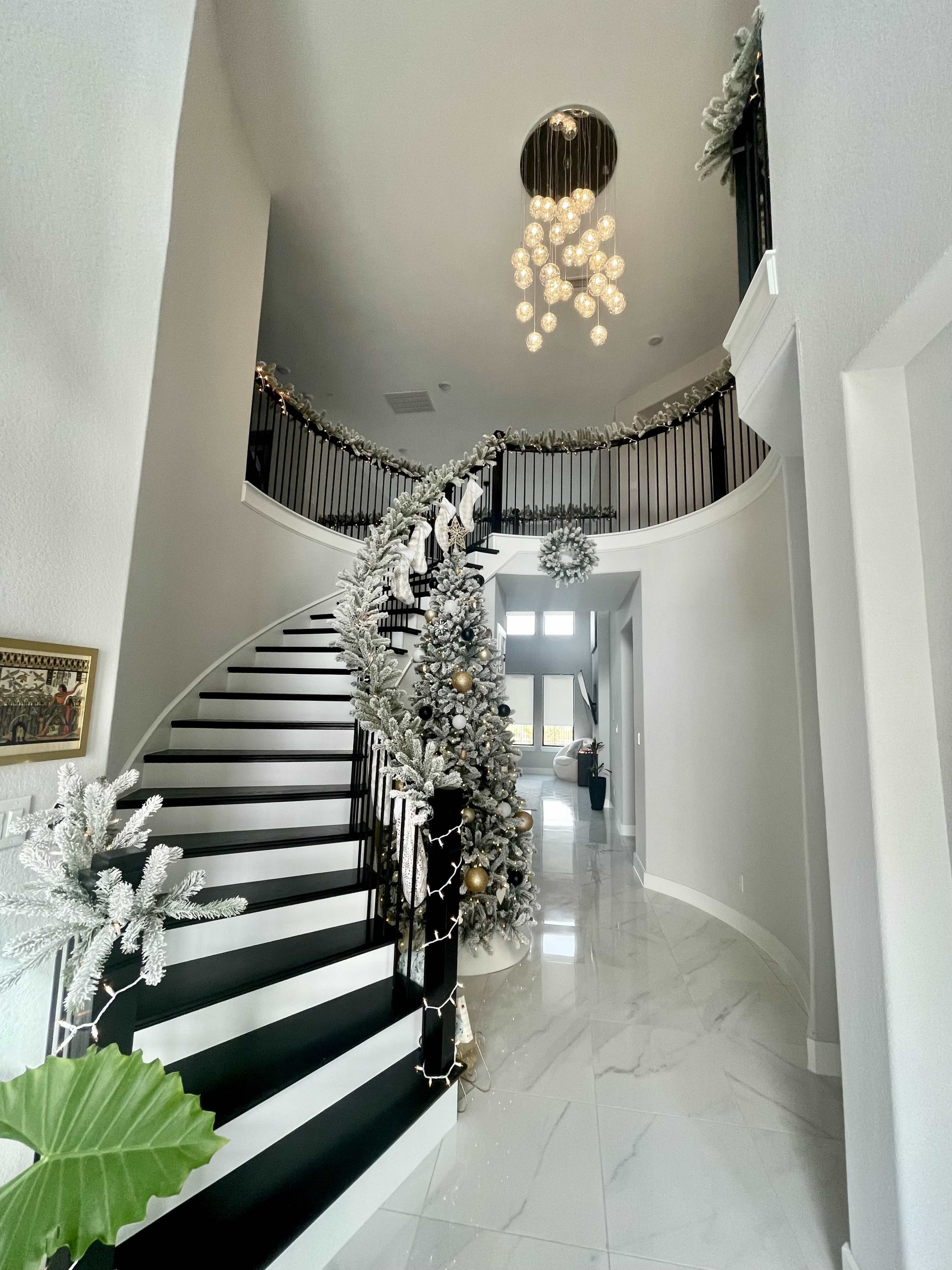 A decorated double staircase with white railings and a chandelier is seen in a bright entryway adorned with holiday decorations.