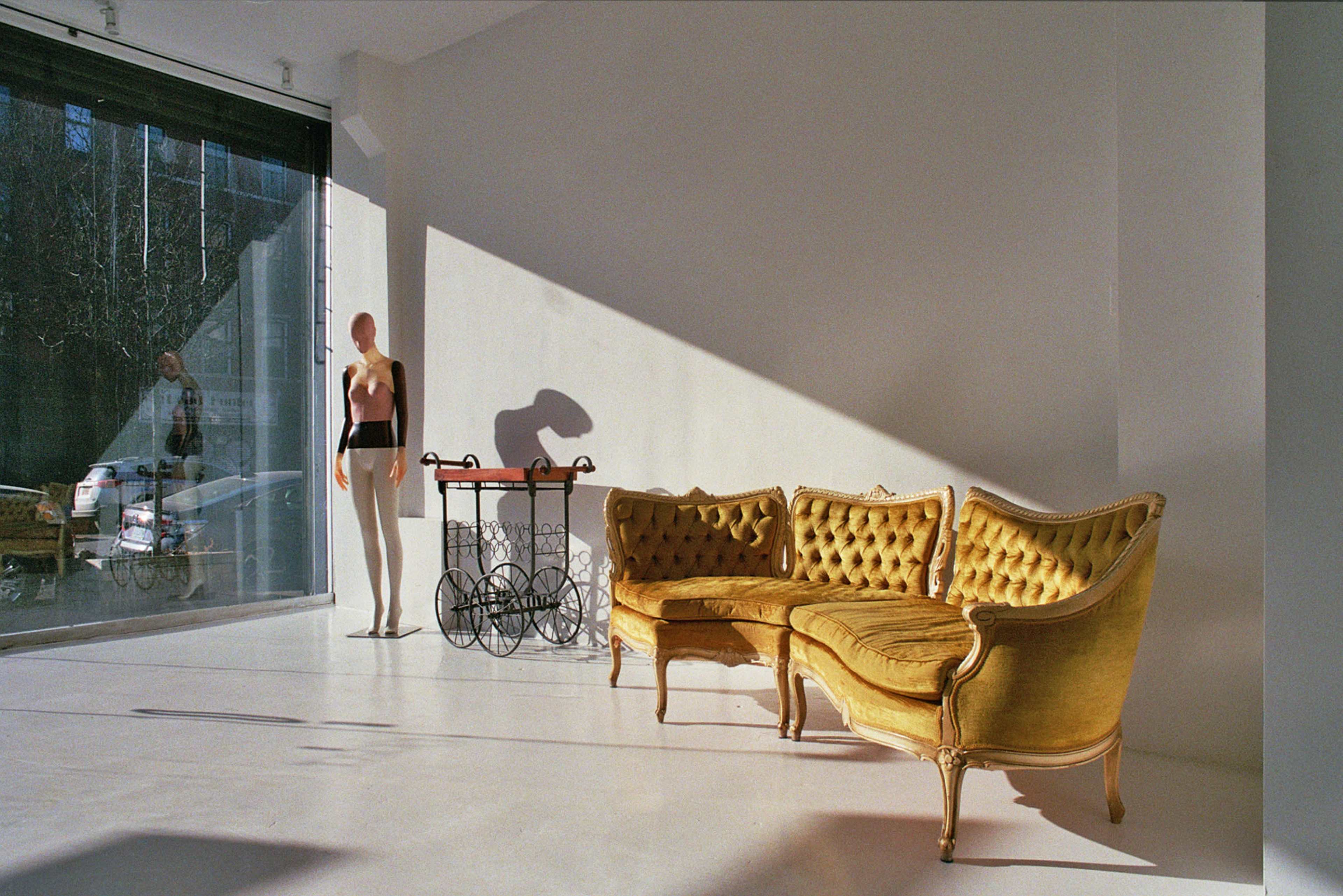 A vintage yellow sofa is positioned beside a large window, with a mannequin standing nearby and a decorative cart in the background.