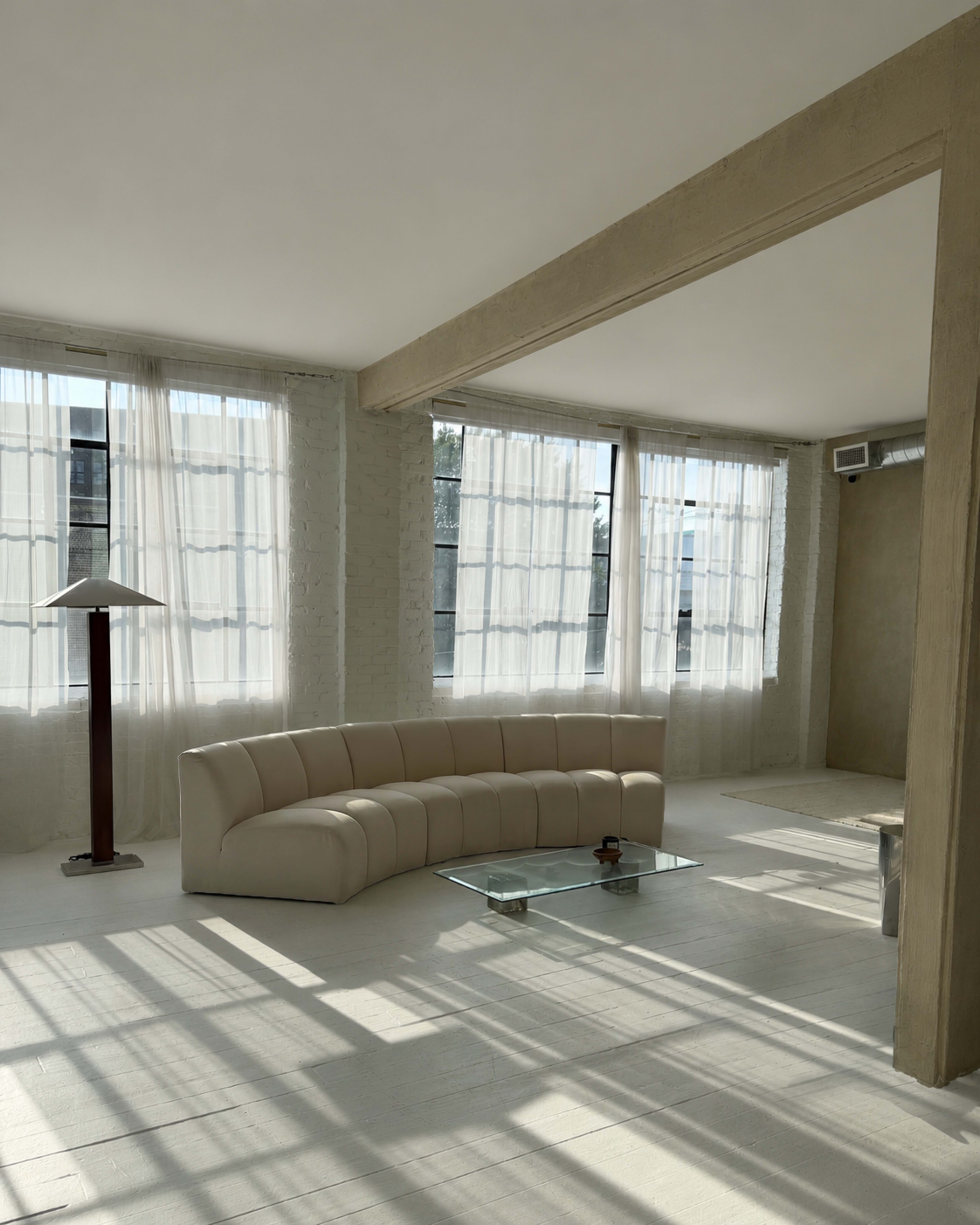 The image shows a modern, minimalist room with a curved white sofa, a glass coffee table, and large windows allowing natural light to flood the space.