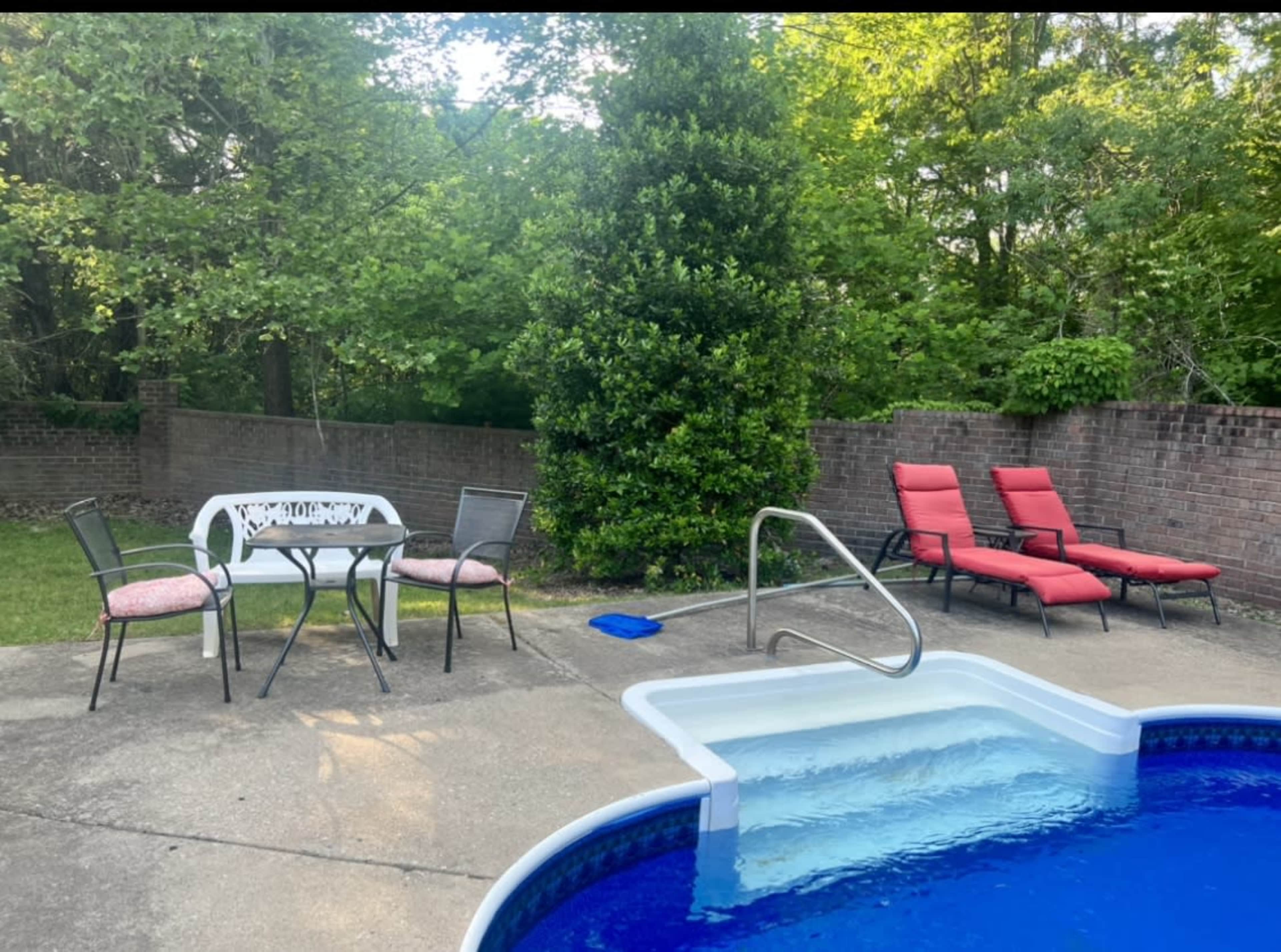 A swimming pool surrounded by two red lounge chairs, a small table with two chairs, and a landscaped area with trees in the background.