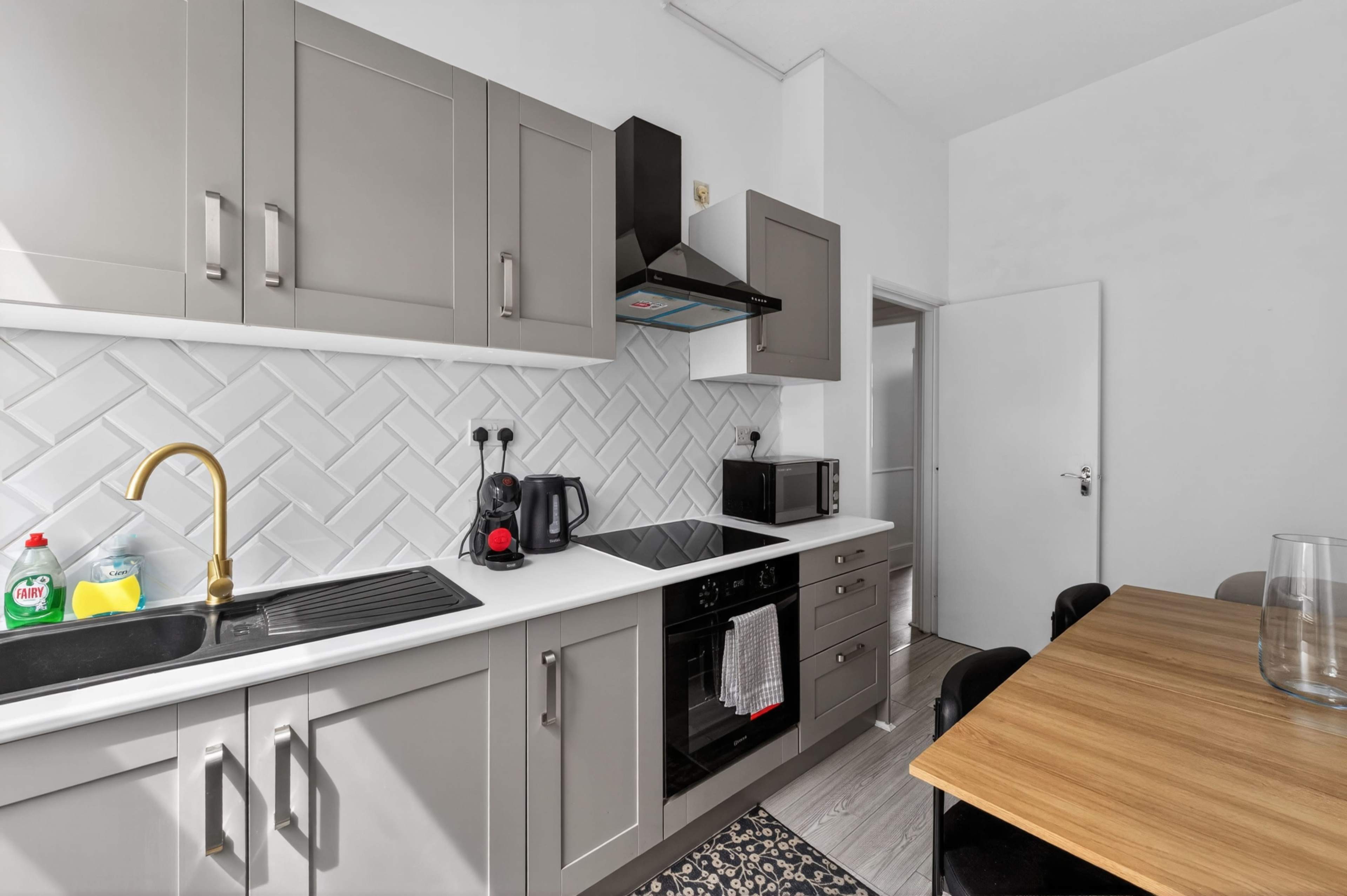 A modern kitchen features gray cabinets, a black oven, a microwave, and a dining table against a light-colored wall with textured backsplash tiles.