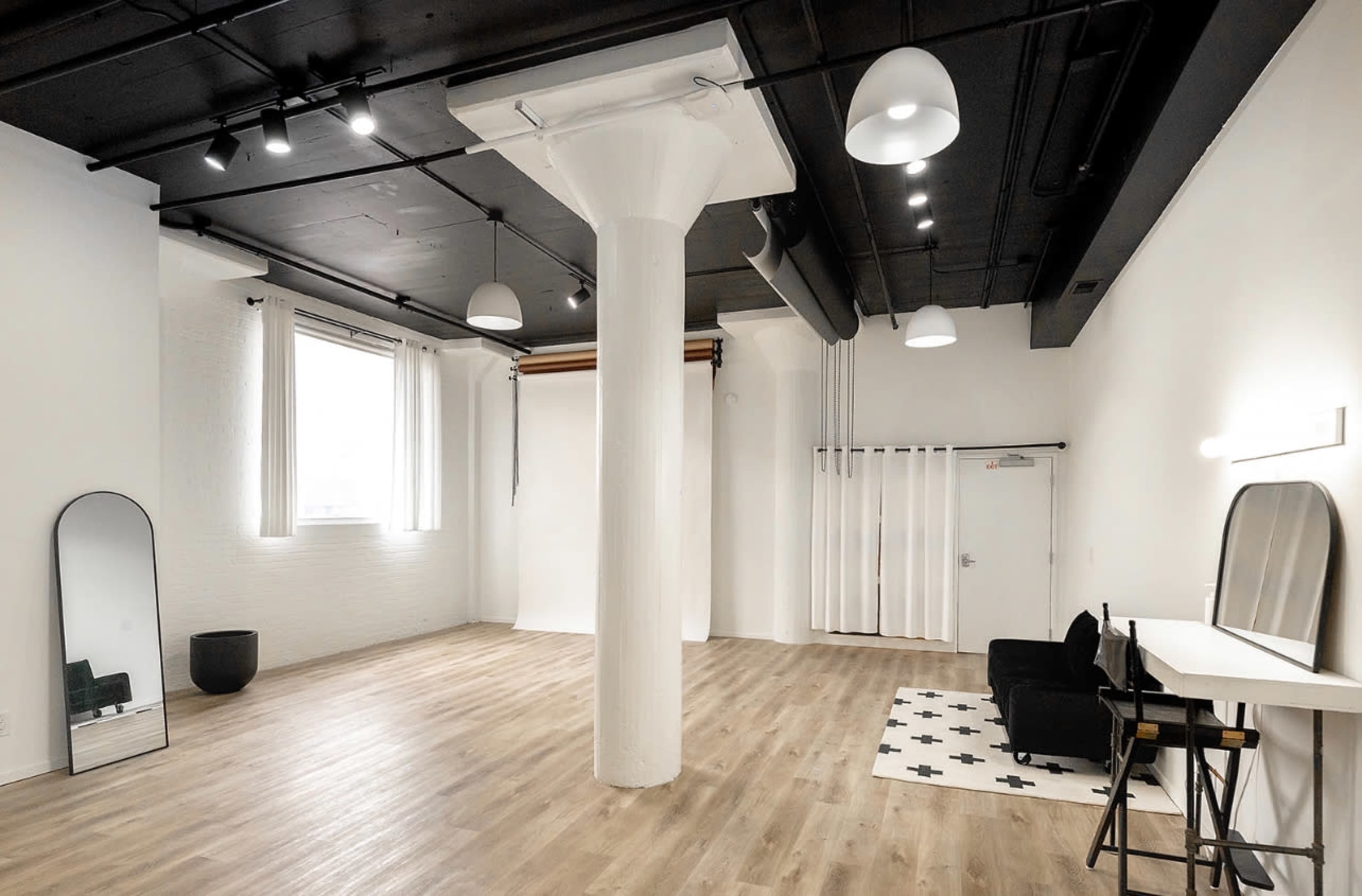 A spacious, minimalist room featuring a central white column, wooden flooring, a large mirror, and simple furniture against a white backdrop.