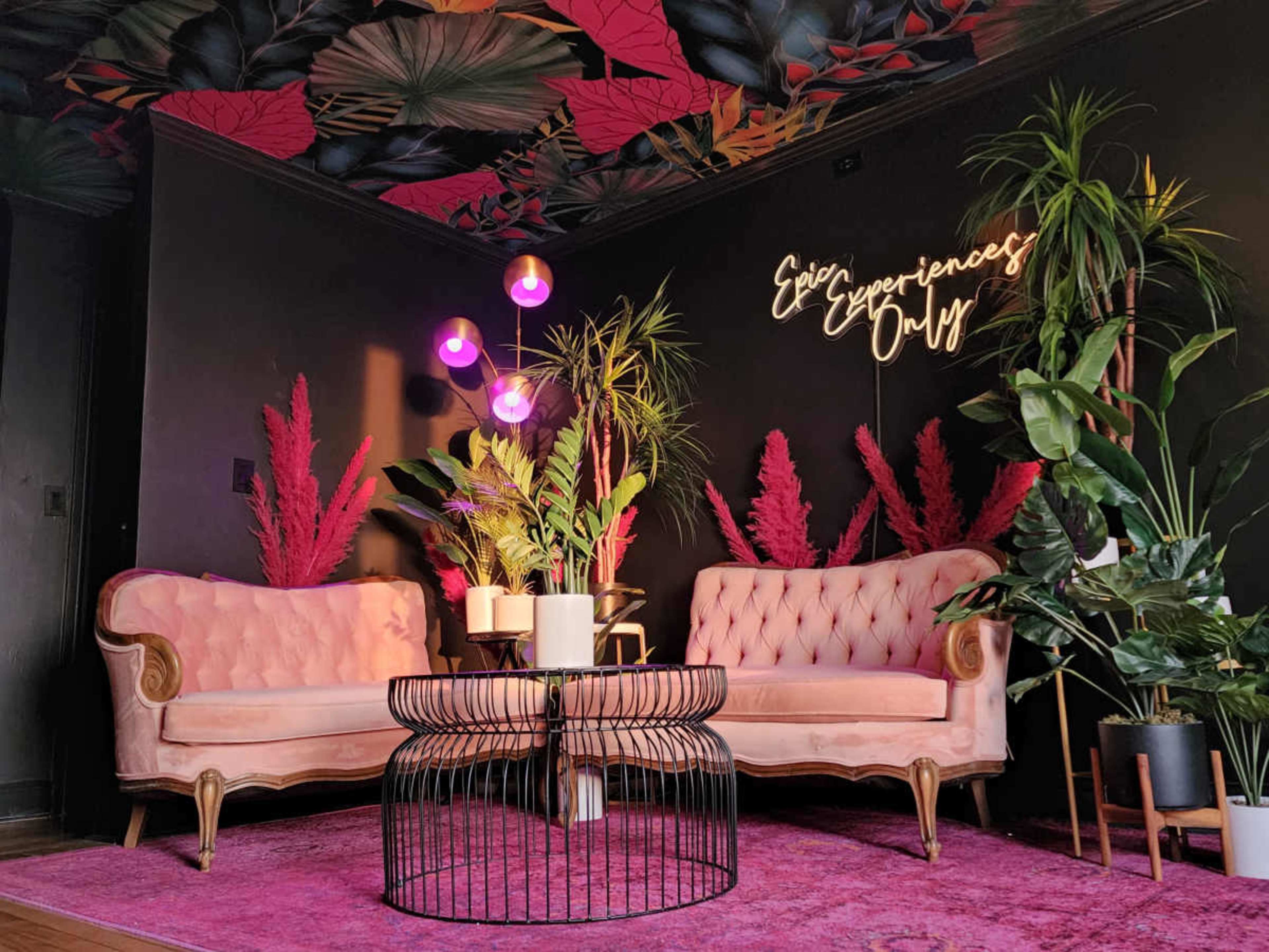A pink and black lounge area features two velvet sofas, a round black metal coffee table, and vibrant plants against a decorative wall with the phrase "Epic Experiences Only."