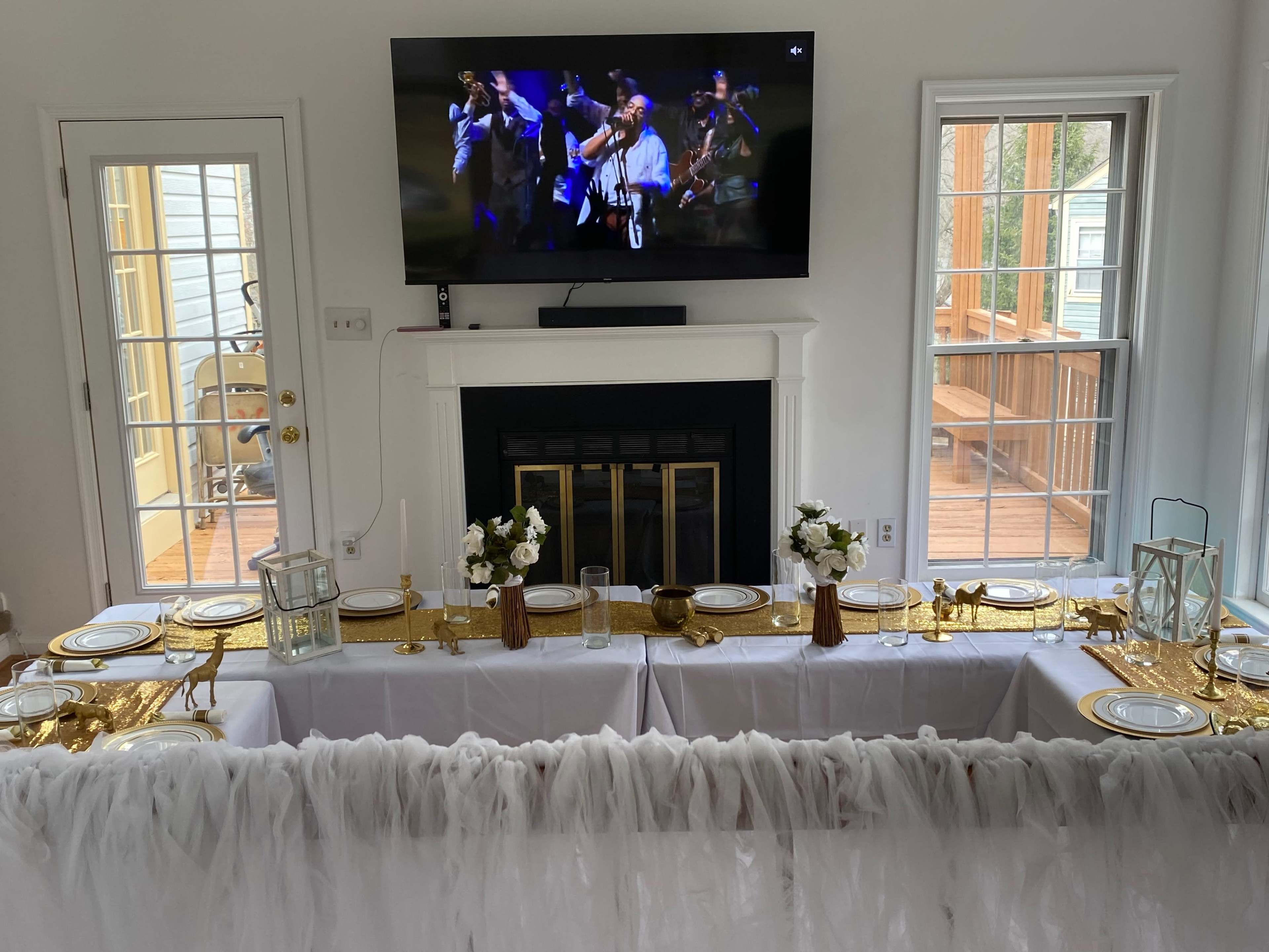 A long dining table is set with white tablecloths and gold accents, with a television displaying a performance mounted above the fireplace.