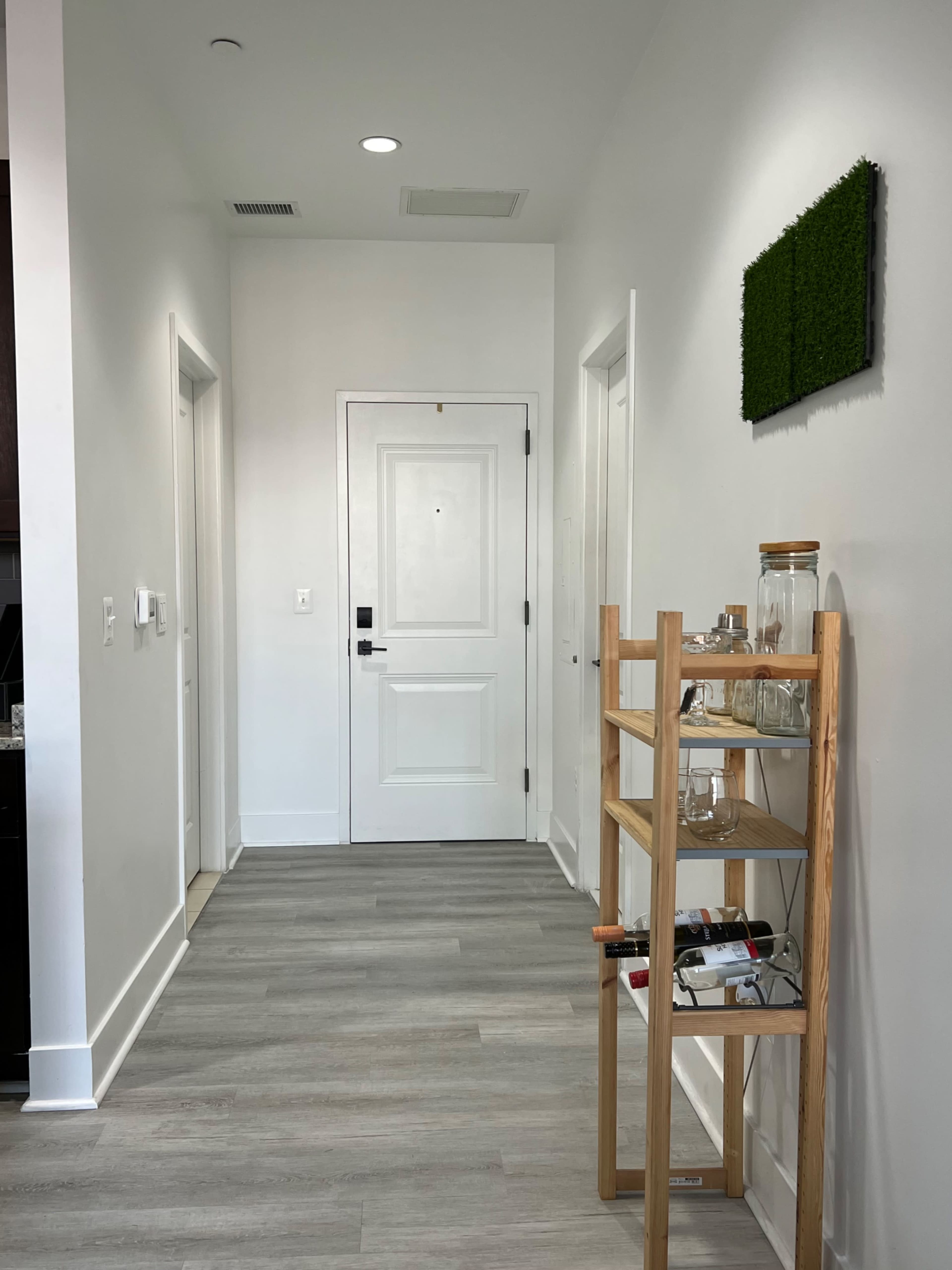 The image shows a narrow hallway leading to a front door, with a wooden shelf holding glass jars on the right and a decorative green wall piece above it.