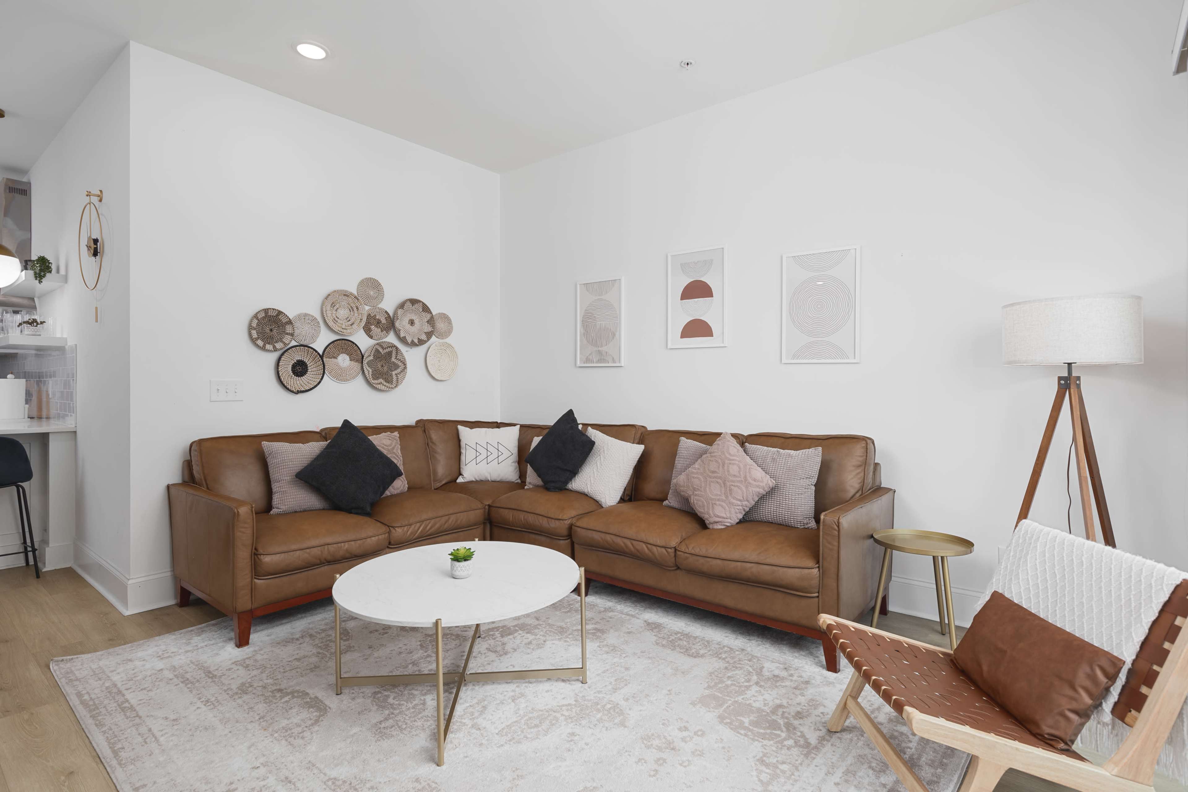 A modern living room features a brown sectional sofa with decorative cushions, a round coffee table, a floor lamp, and framed artwork on the walls.