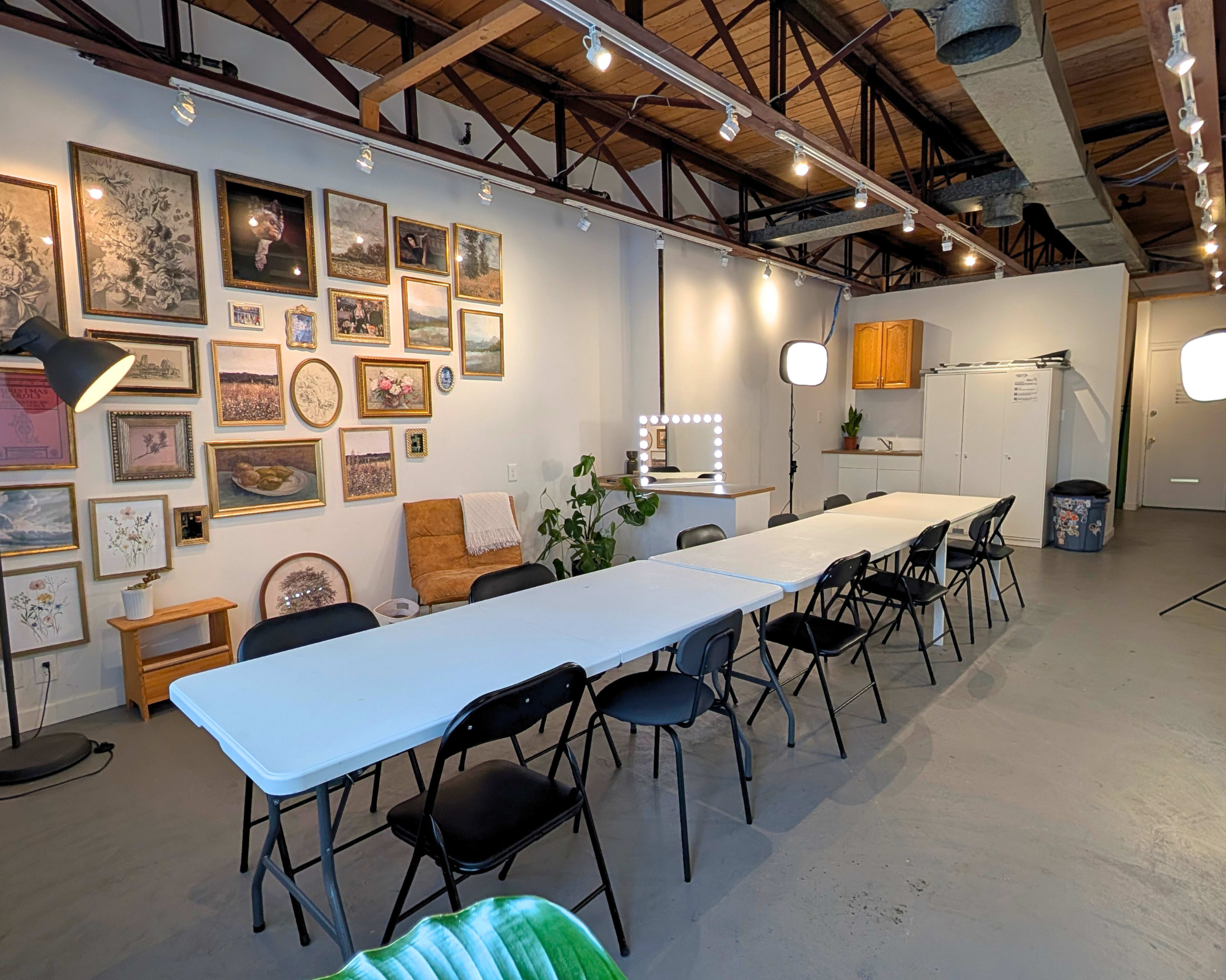 The image shows a spacious room with a long table surrounded by chairs, decorated with various framed pictures on the wall, and featuring a lighted makeup station in the corner.
