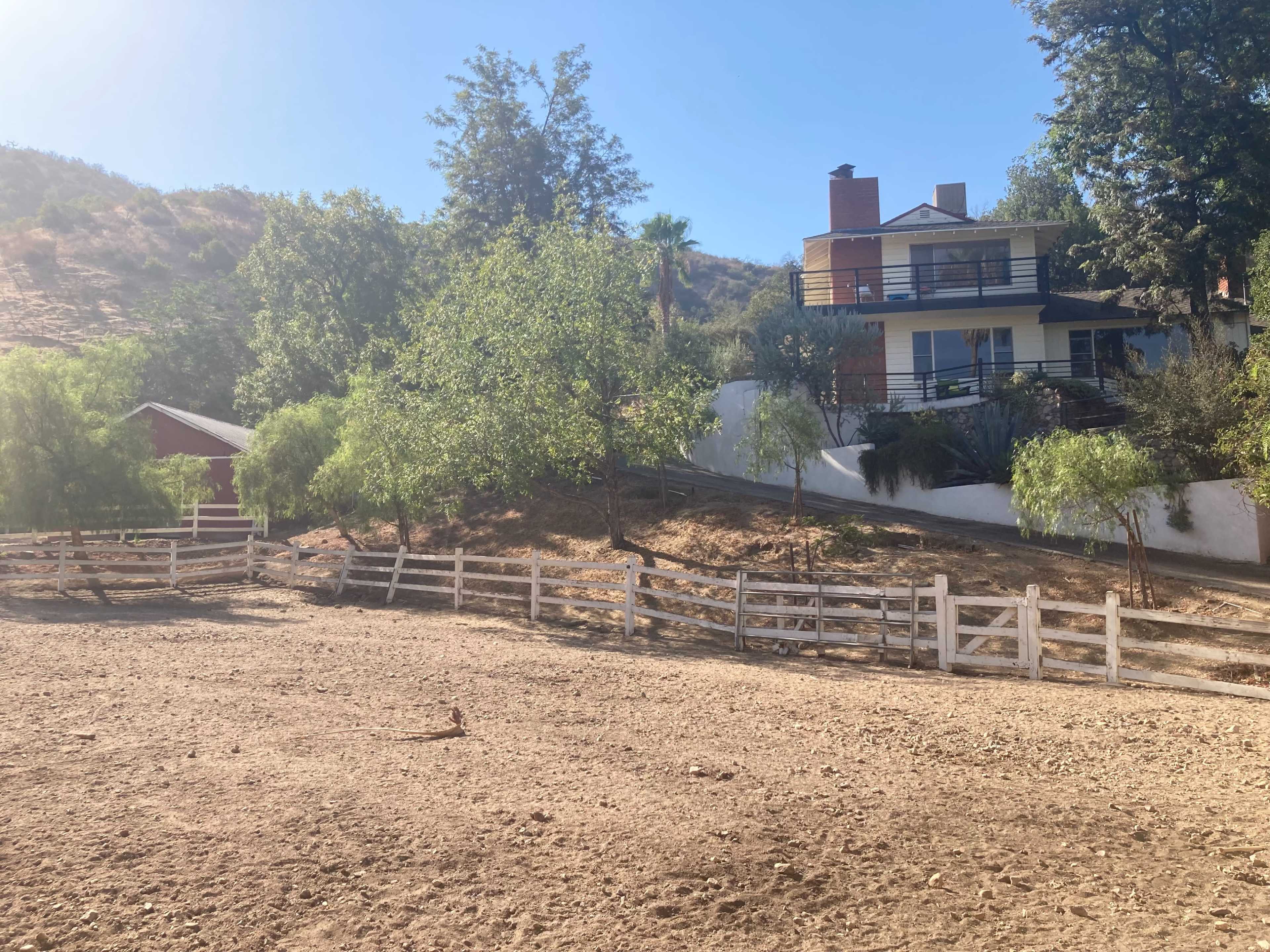 A modern house with a deck overlooks a fenced area and a red barn on a hillside.