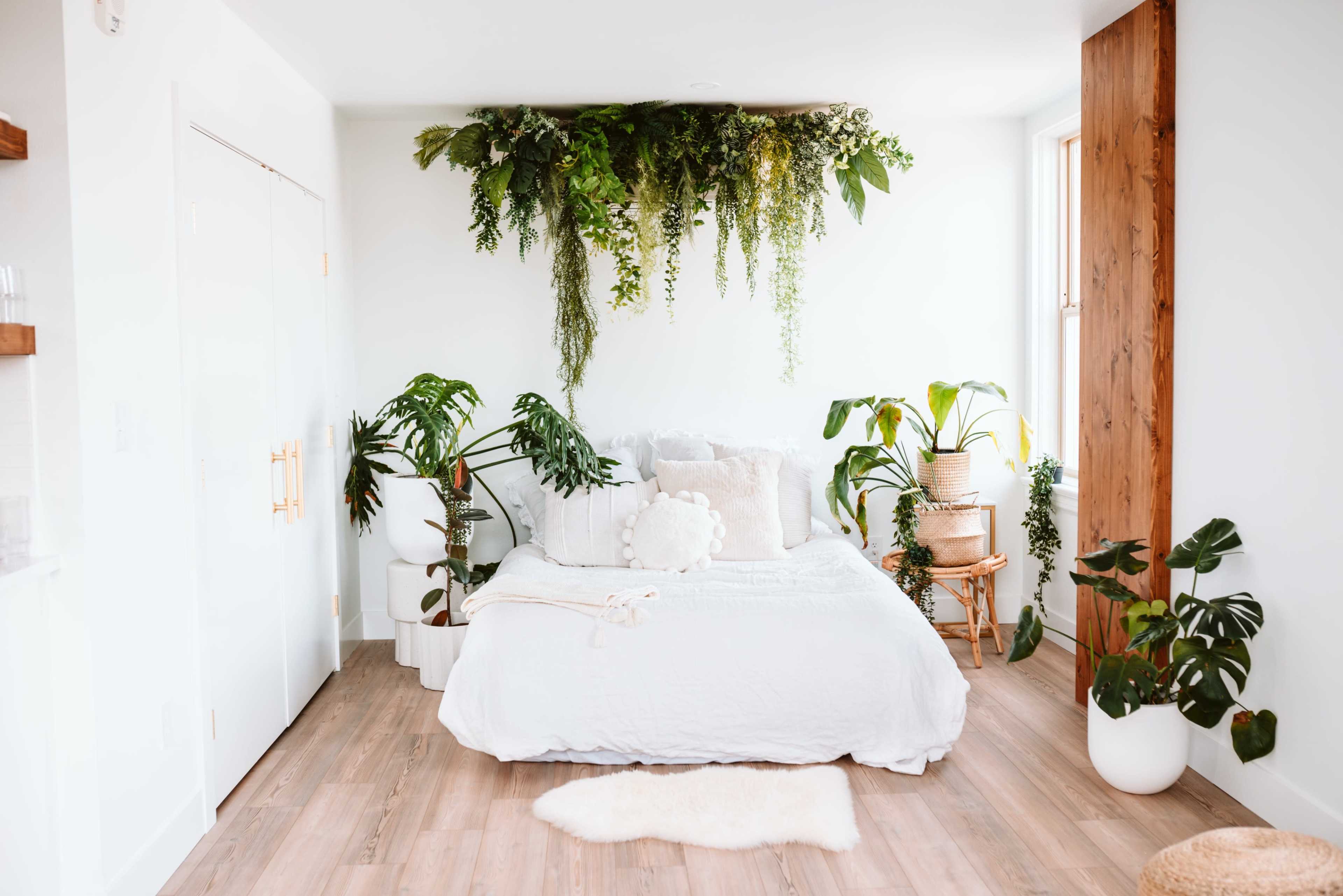 The image shows a bright bedroom featuring a white bed adorned with pillows, surrounded by various indoor plants and wooden accents.