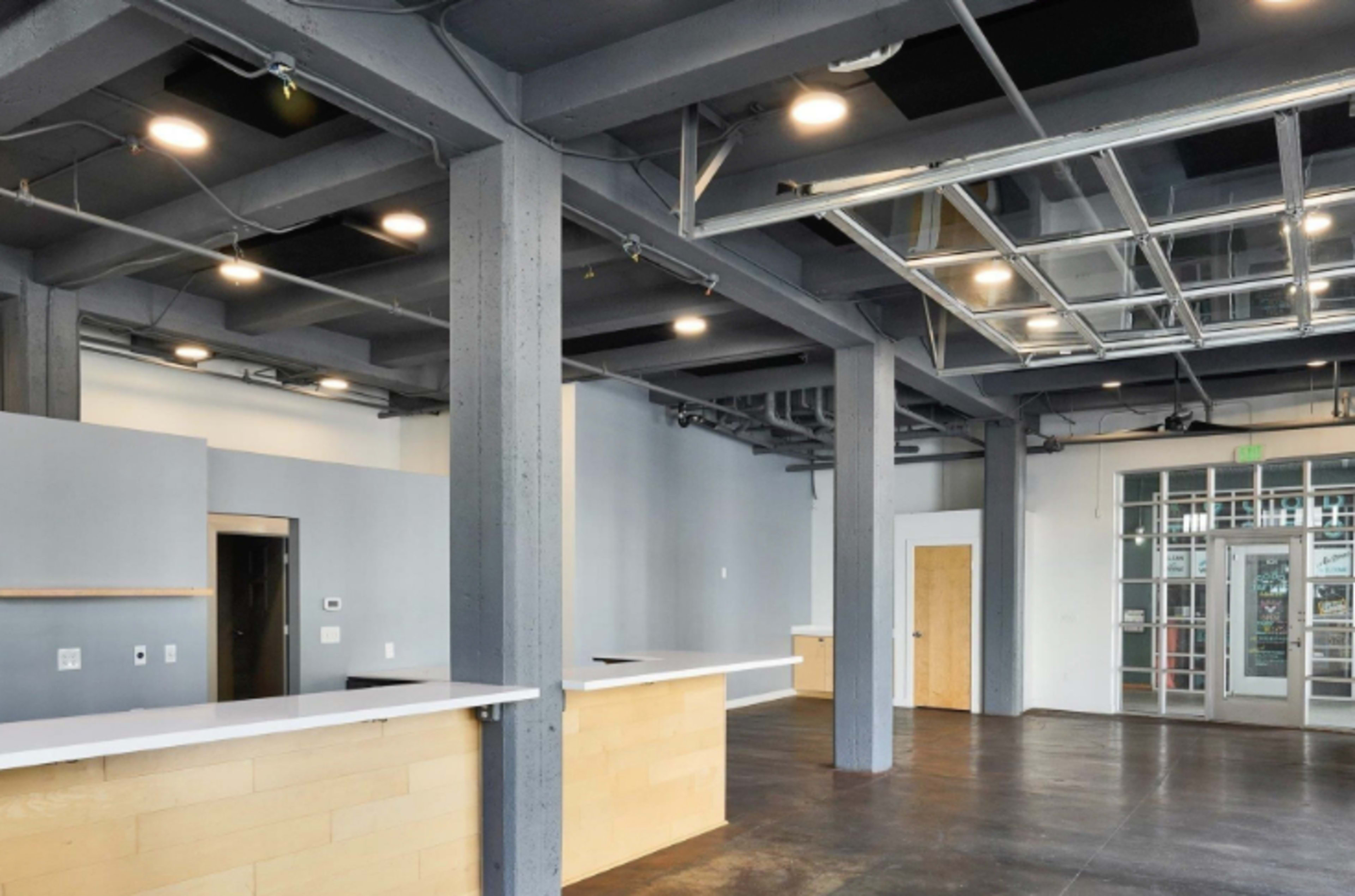 The image shows an empty commercial space with grey concrete columns, high ceilings, and a reception area featuring white countertops and a glass front entrance.