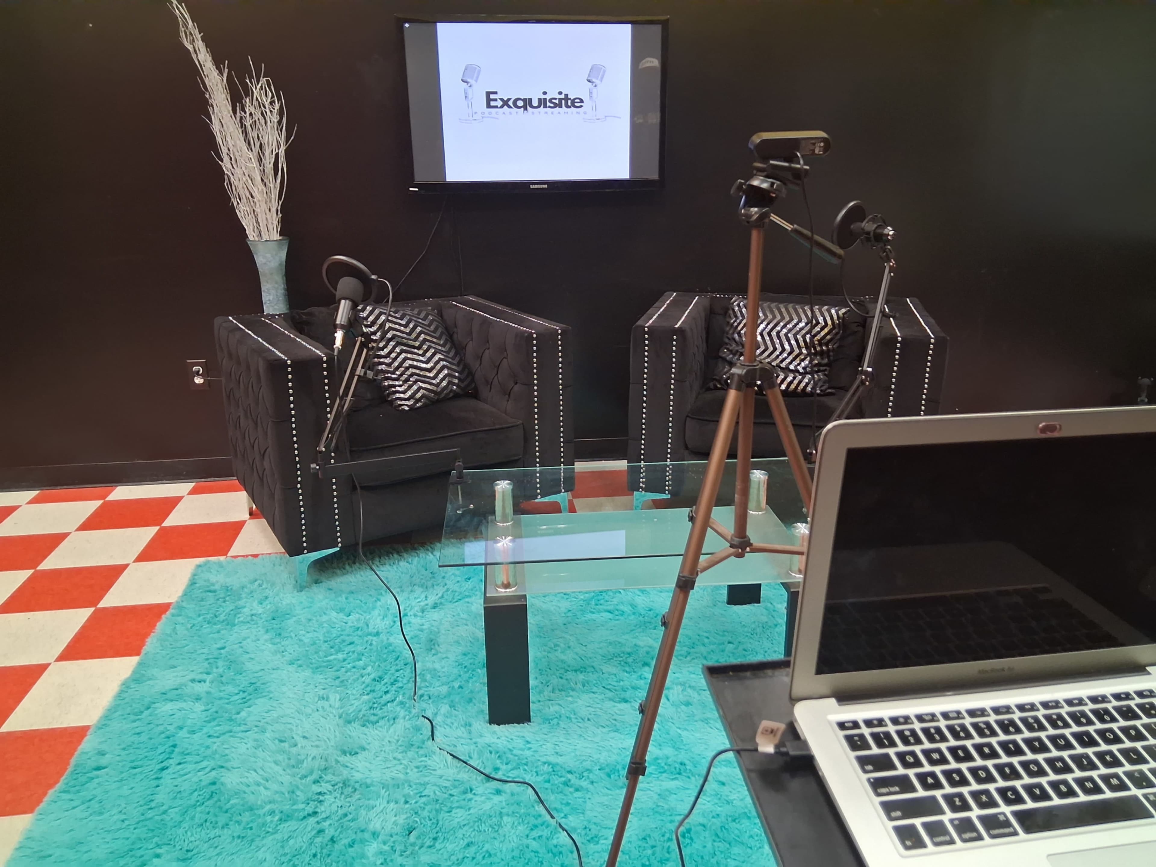 The image shows a recording studio setup featuring two black armchairs with decorative pillows, a glass coffee table, a blue rug, and a television screen displaying the word "Exquisite."