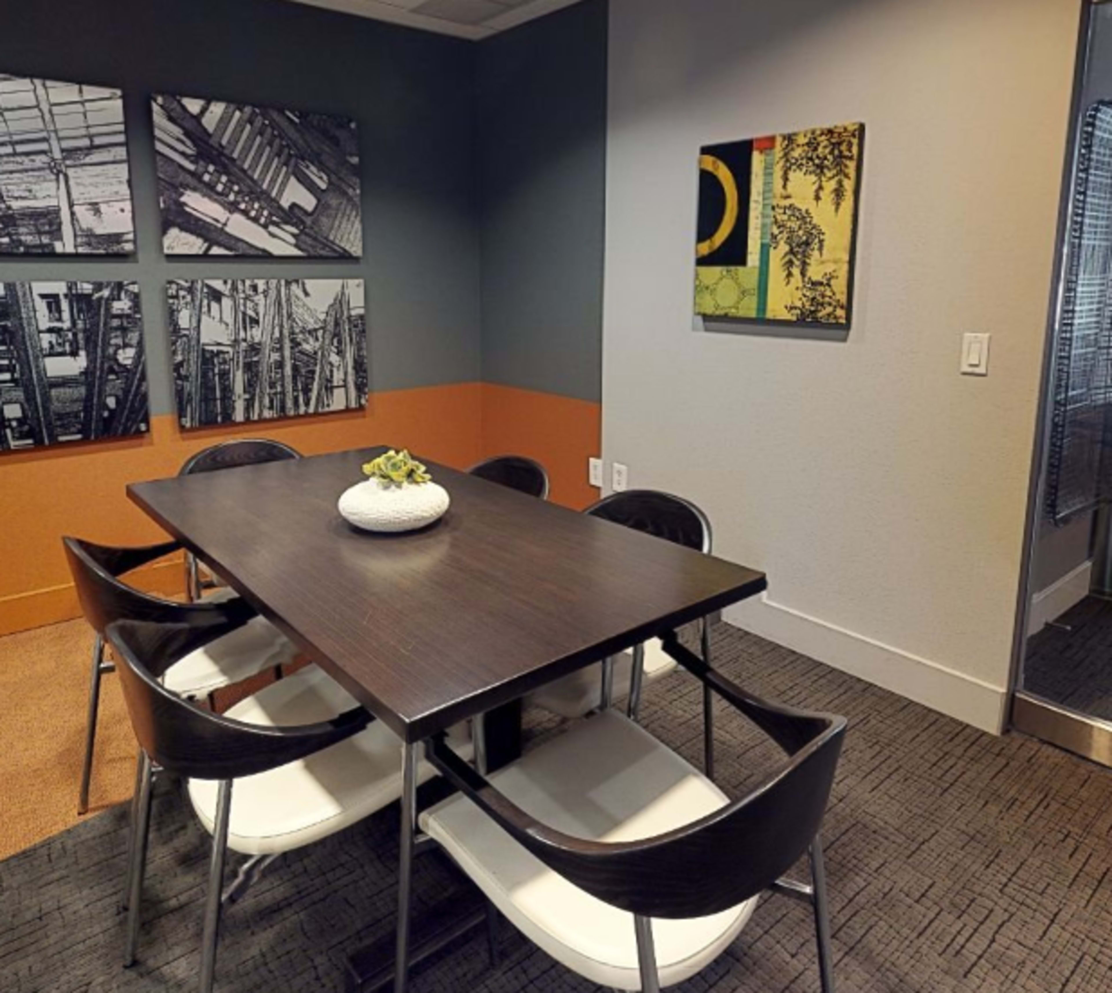 A meeting room furnished with a dark wooden table and six white chairs, accented by framed artworks on the walls and a potted plant as a centerpiece.