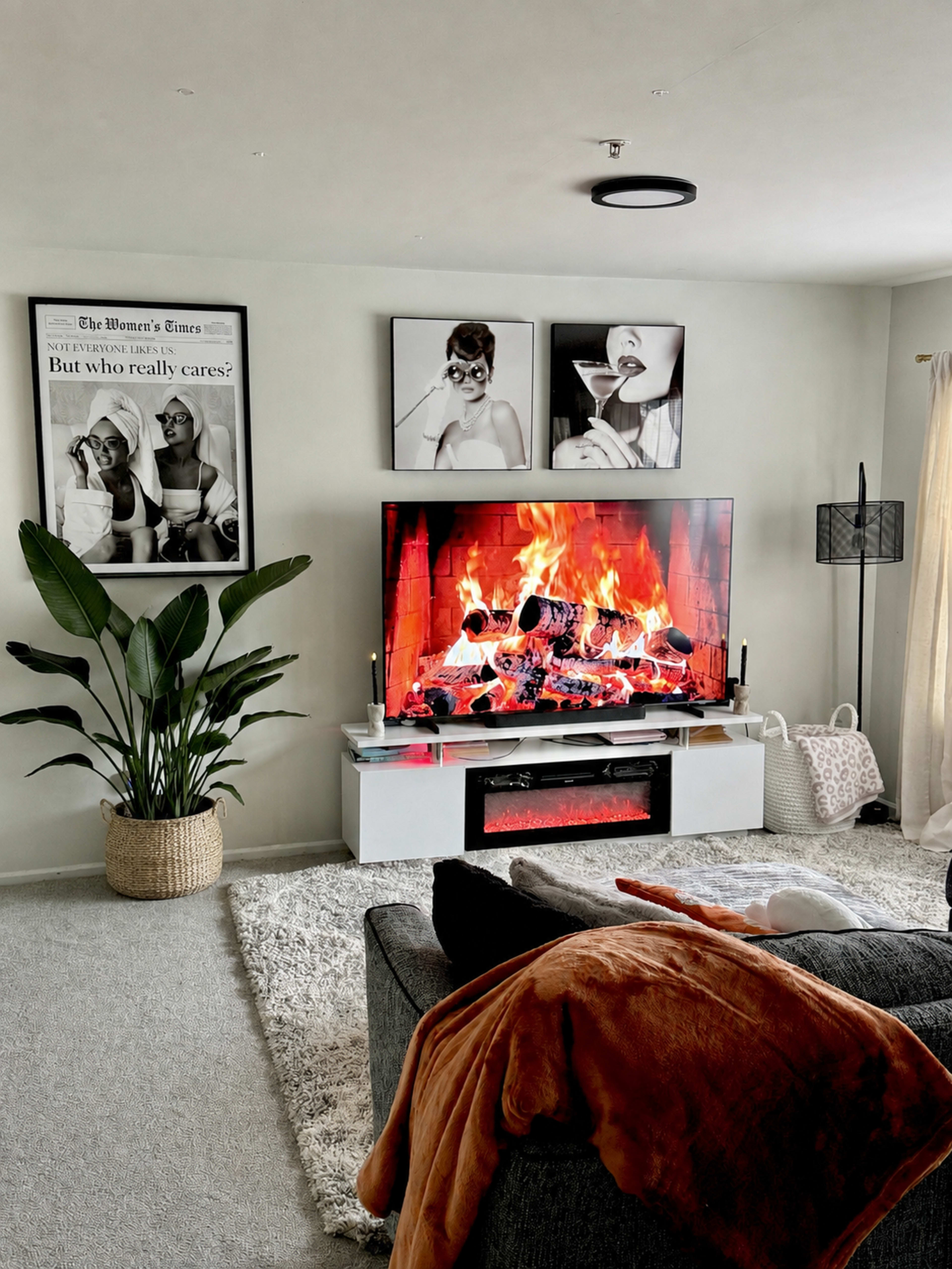 A modern living room features a large television displaying a digital fireplace, surrounded by artwork and a potted plant.