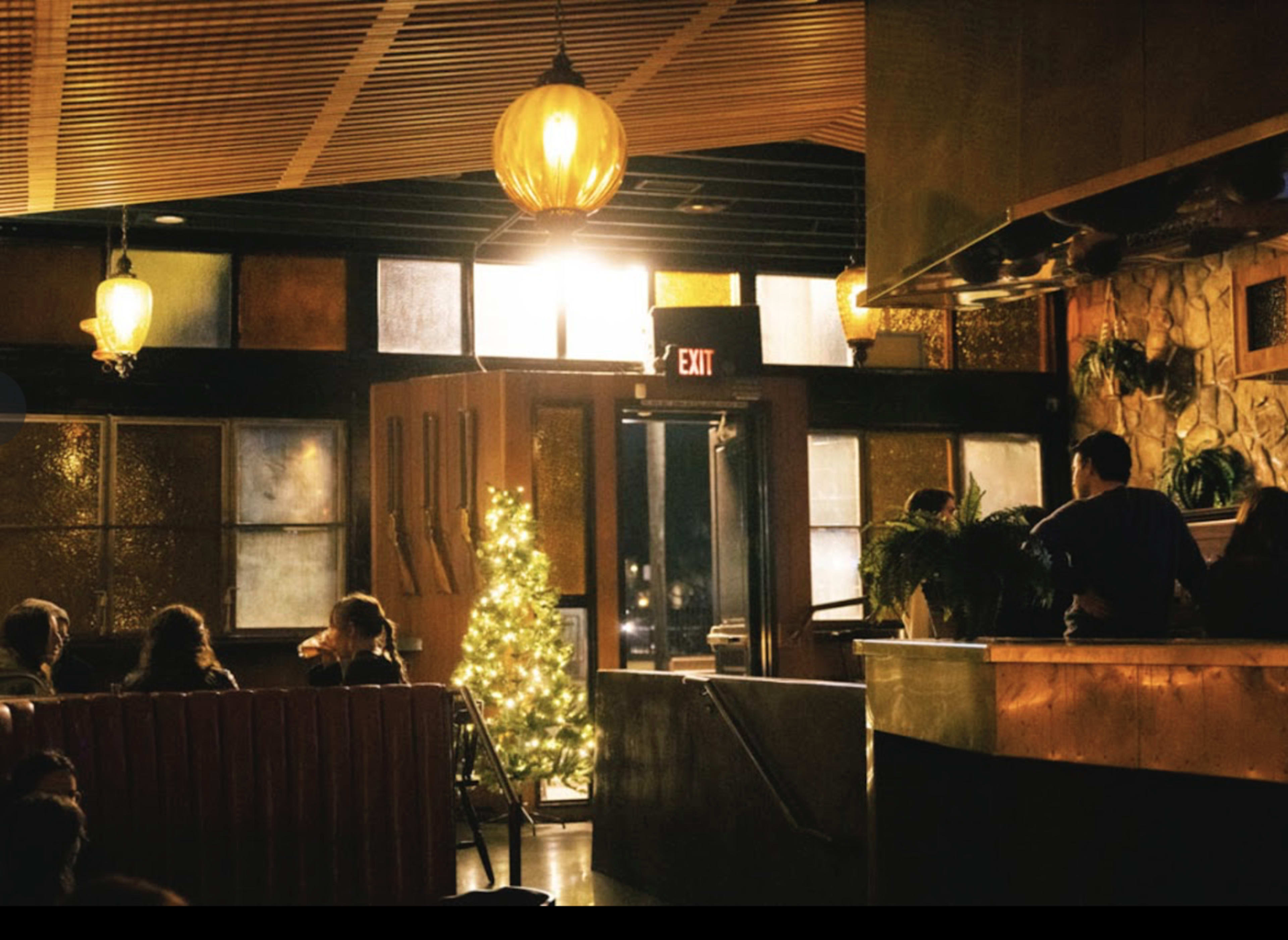 A warmly lit bar features a small, decorated Christmas tree near the entrance, while patrons sit at tables and a bartender stands behind the counter.