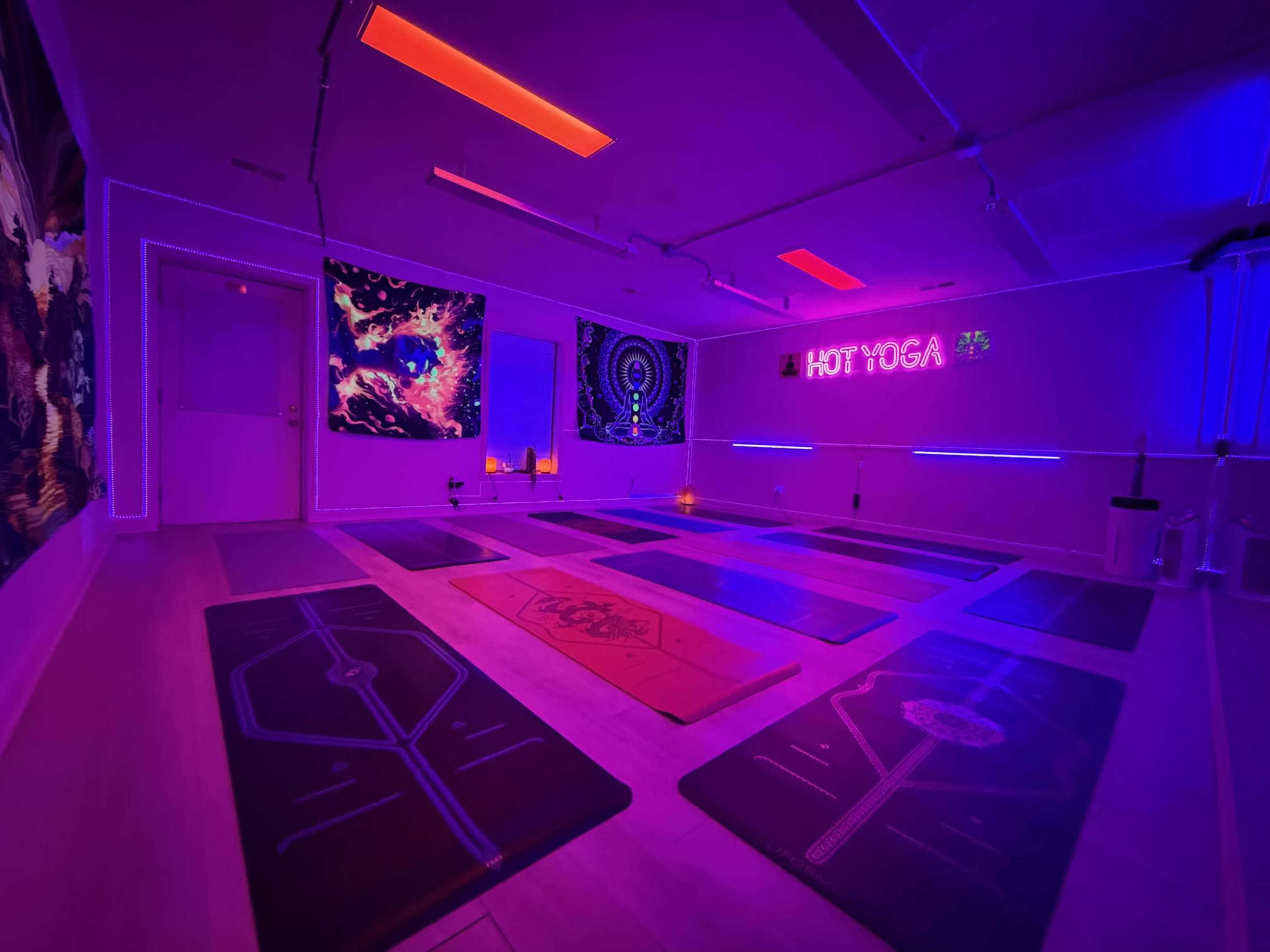 The image shows a dimly lit yoga studio featuring colorful neon lights, yoga mats arranged on the floor, and decorative wall art.