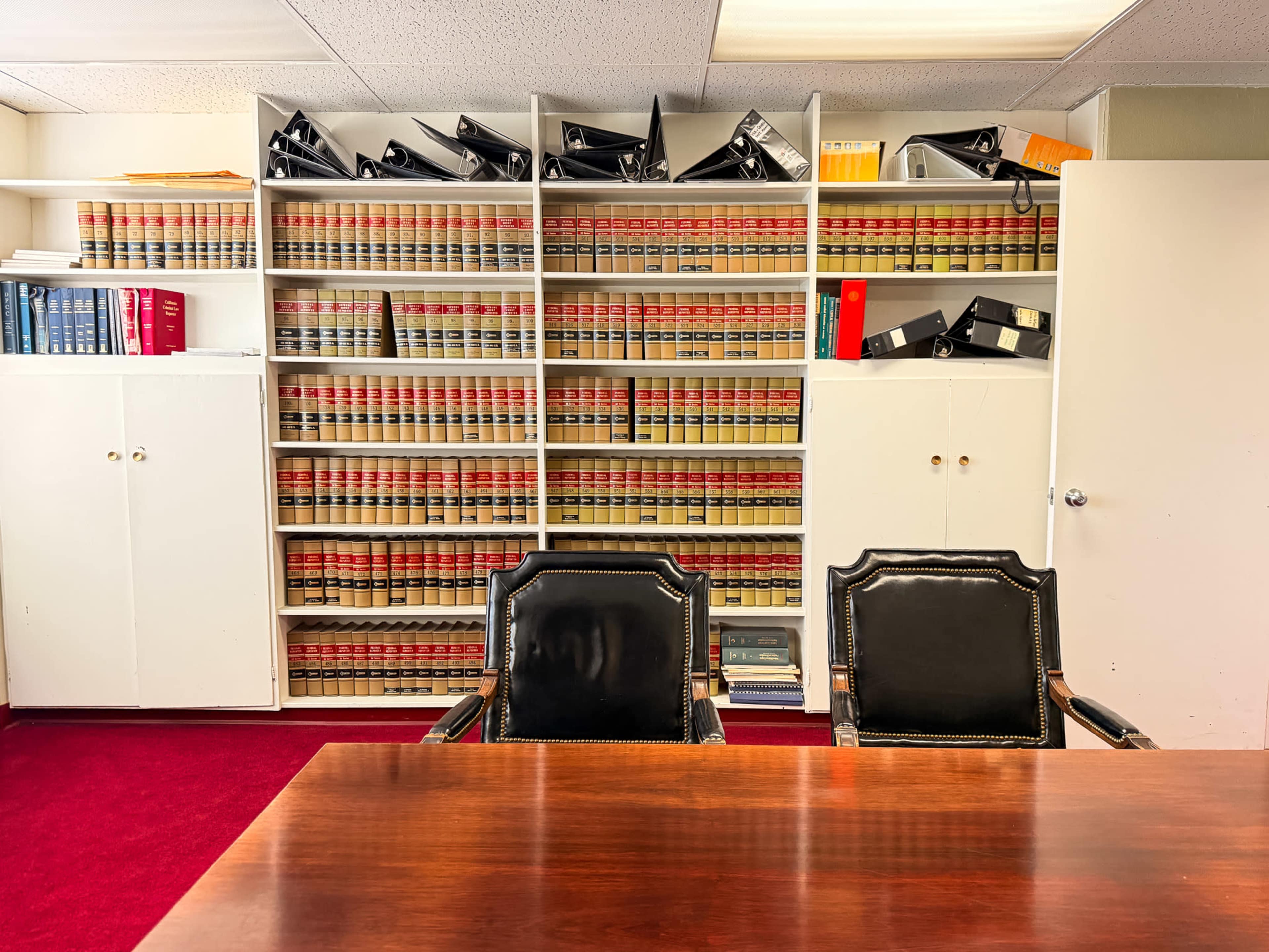 The image shows a small office with a wooden desk and two black chairs facing a large bookshelf filled with legal books and several file boxes on top.