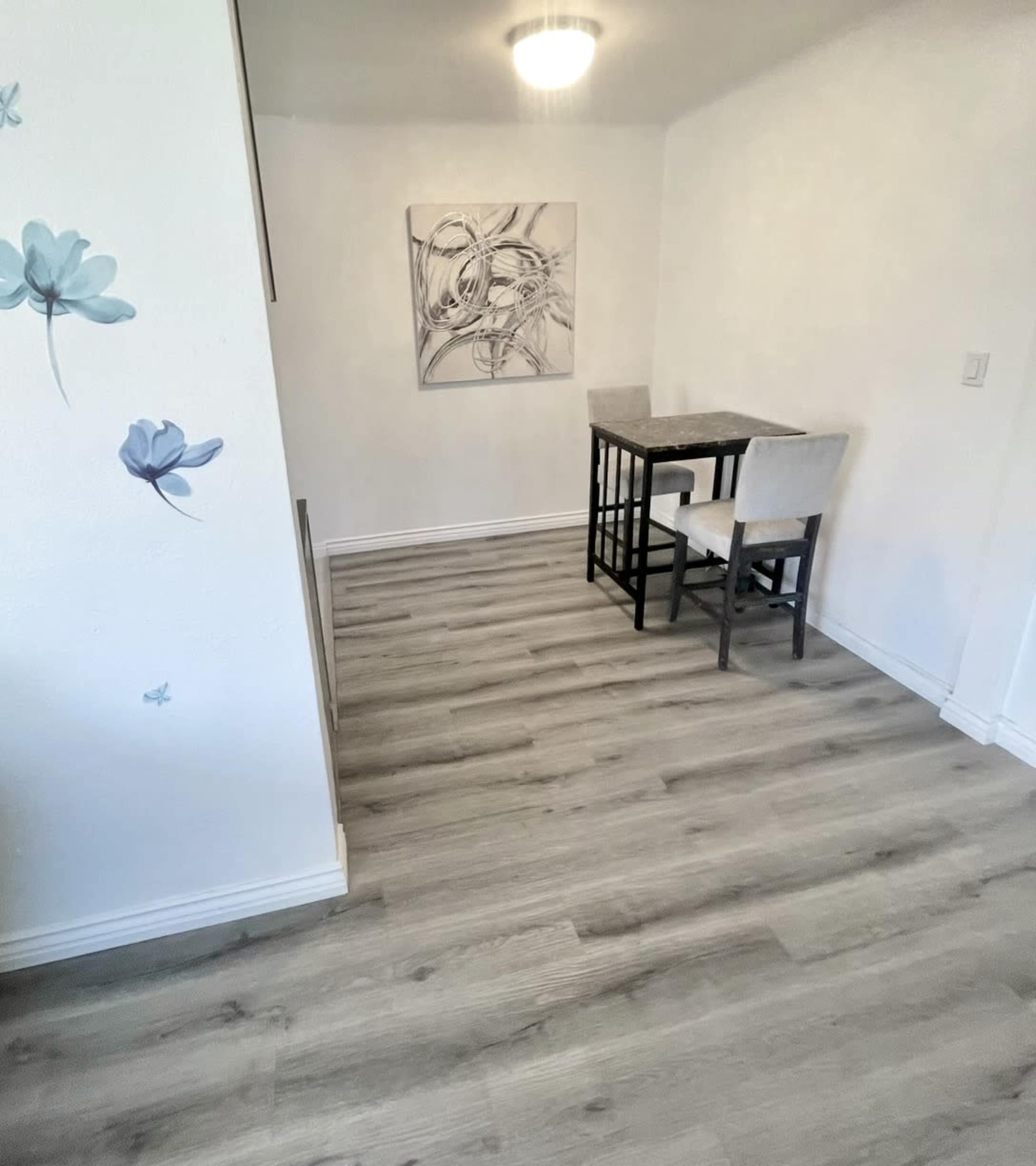 The image shows a light-colored room with wooden flooring, featuring a small table and two chairs along with a wall art piece.