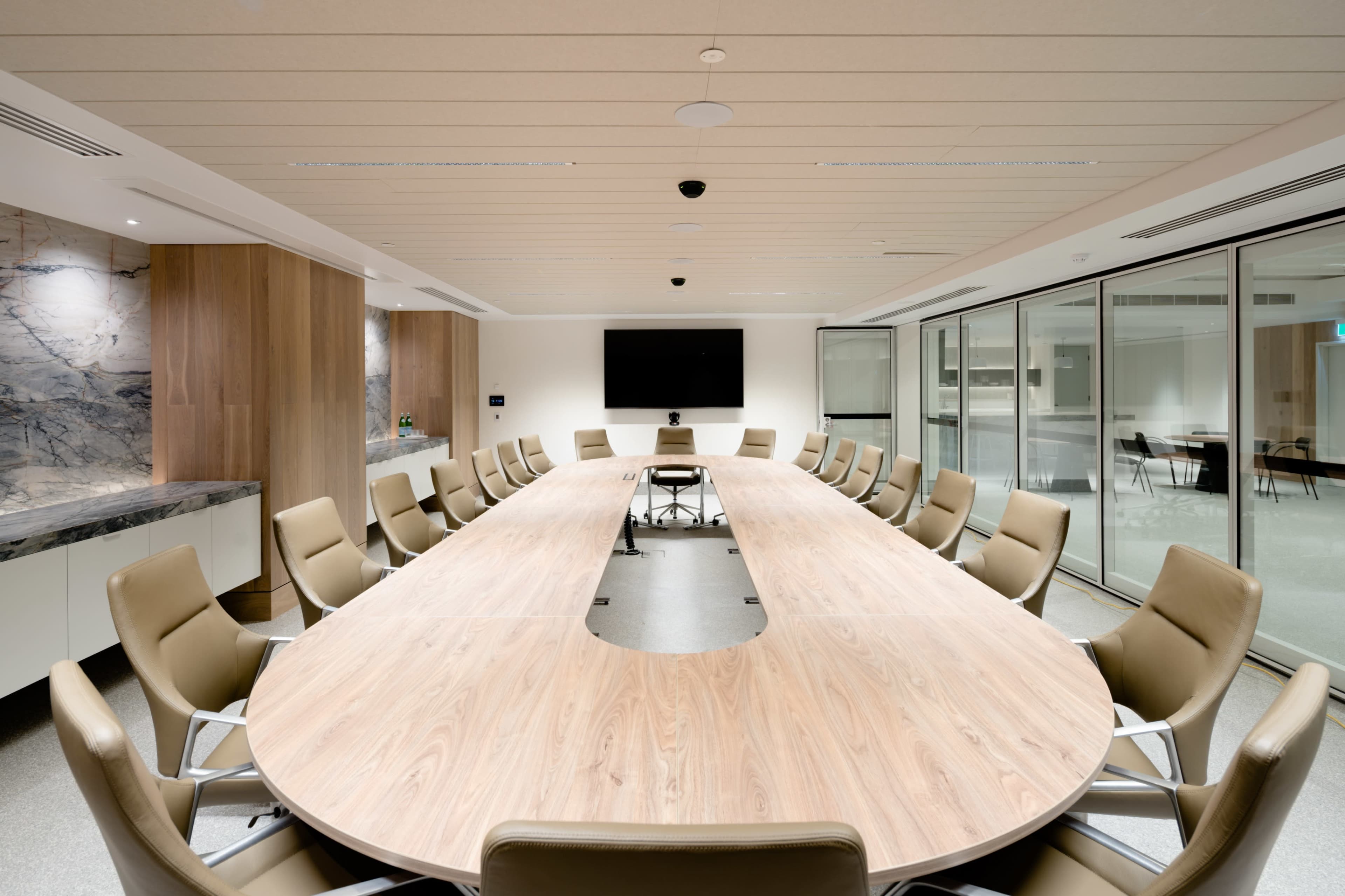 A modern conference room featuring a large oval table surrounded by leather chairs and a wall-mounted screen.