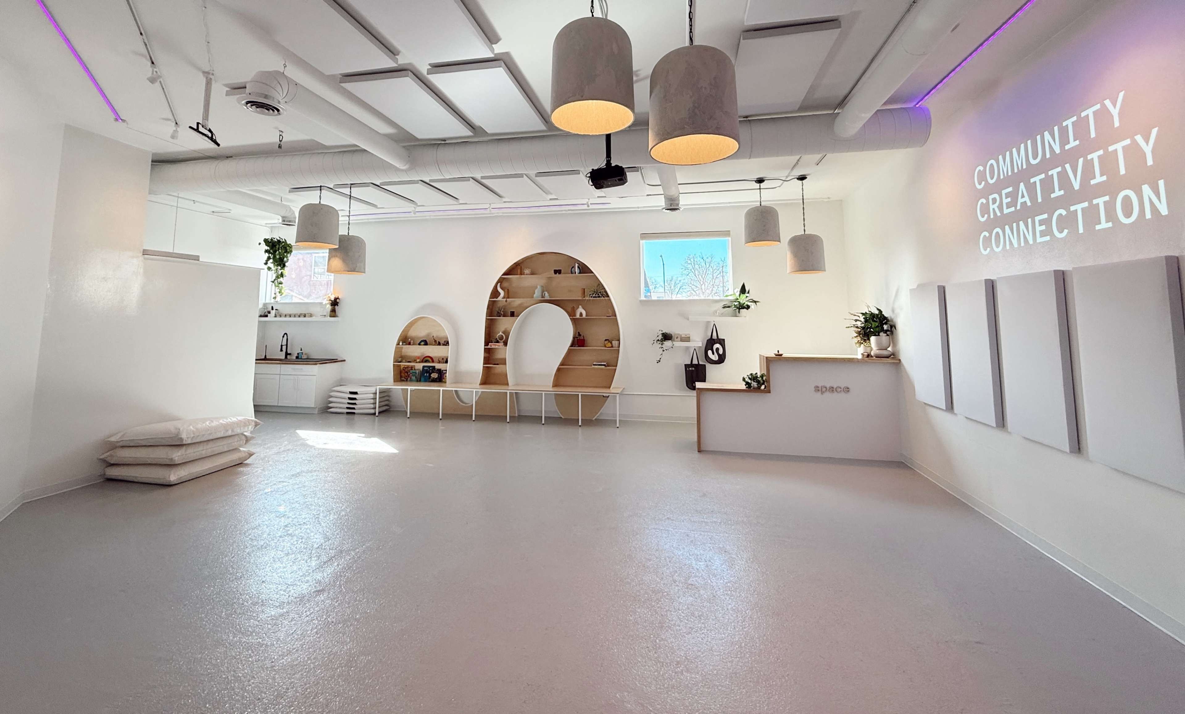 The image shows a bright, minimalistic indoor space featuring a reception area, shelves, and several hanging light fixtures.