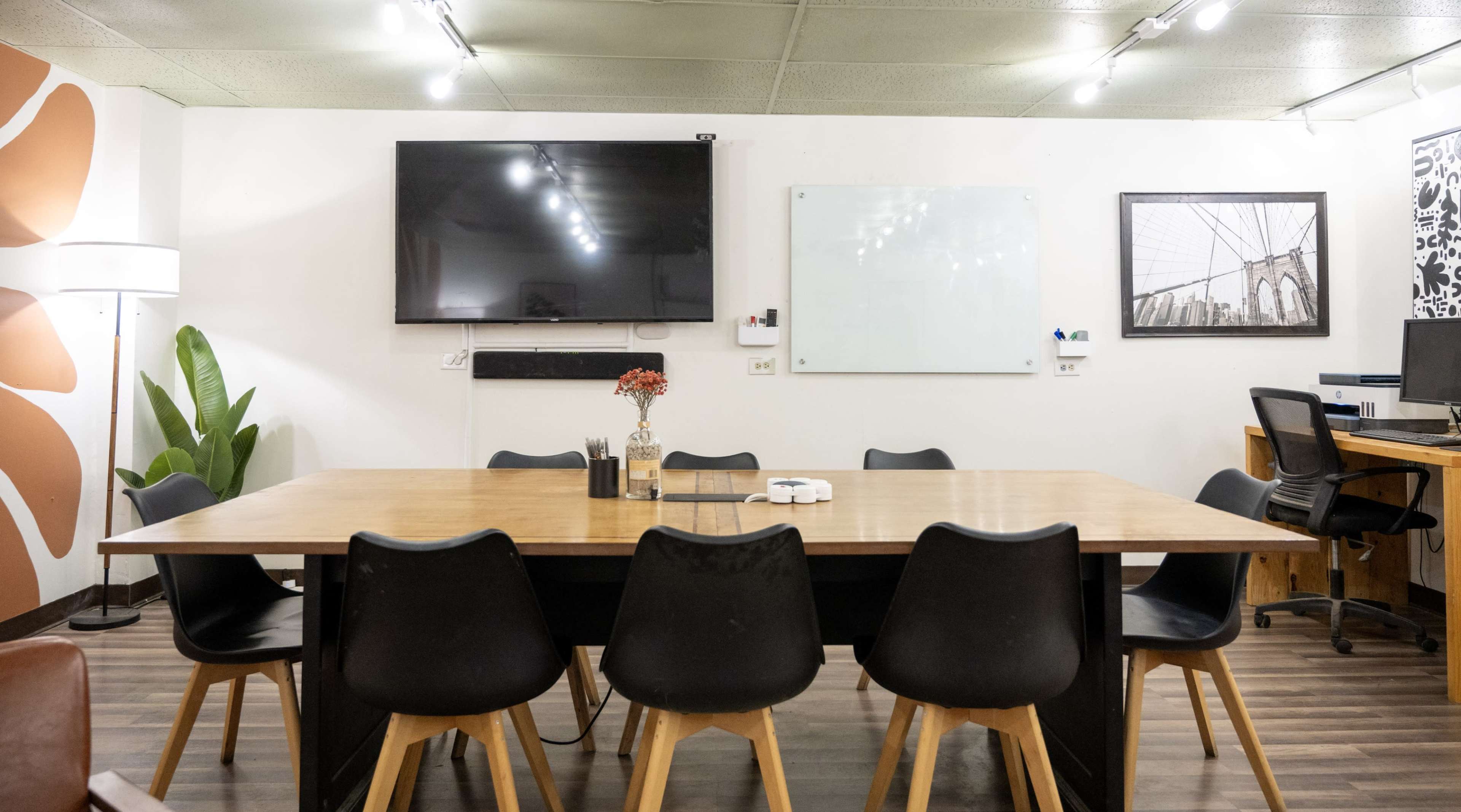 The image shows a modern conference room with a large wooden table surrounded by black chairs, a whiteboard, and a television mounted on the wall.
