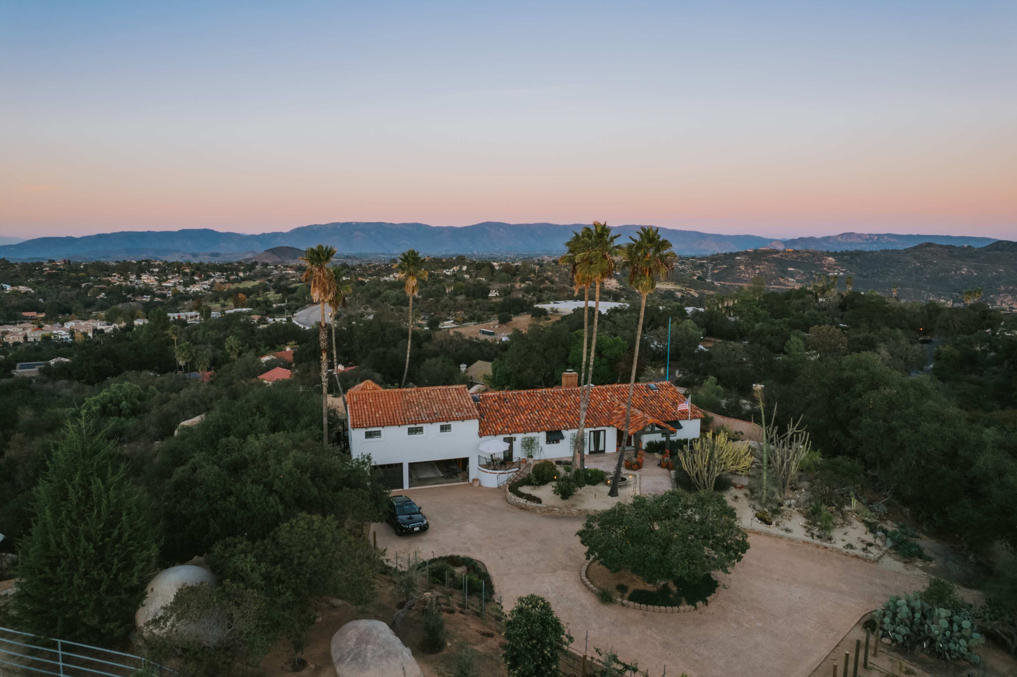 A large house with a red tiled roof is surrounded by palm trees and greenery, set against a hilly landscape at dusk.
