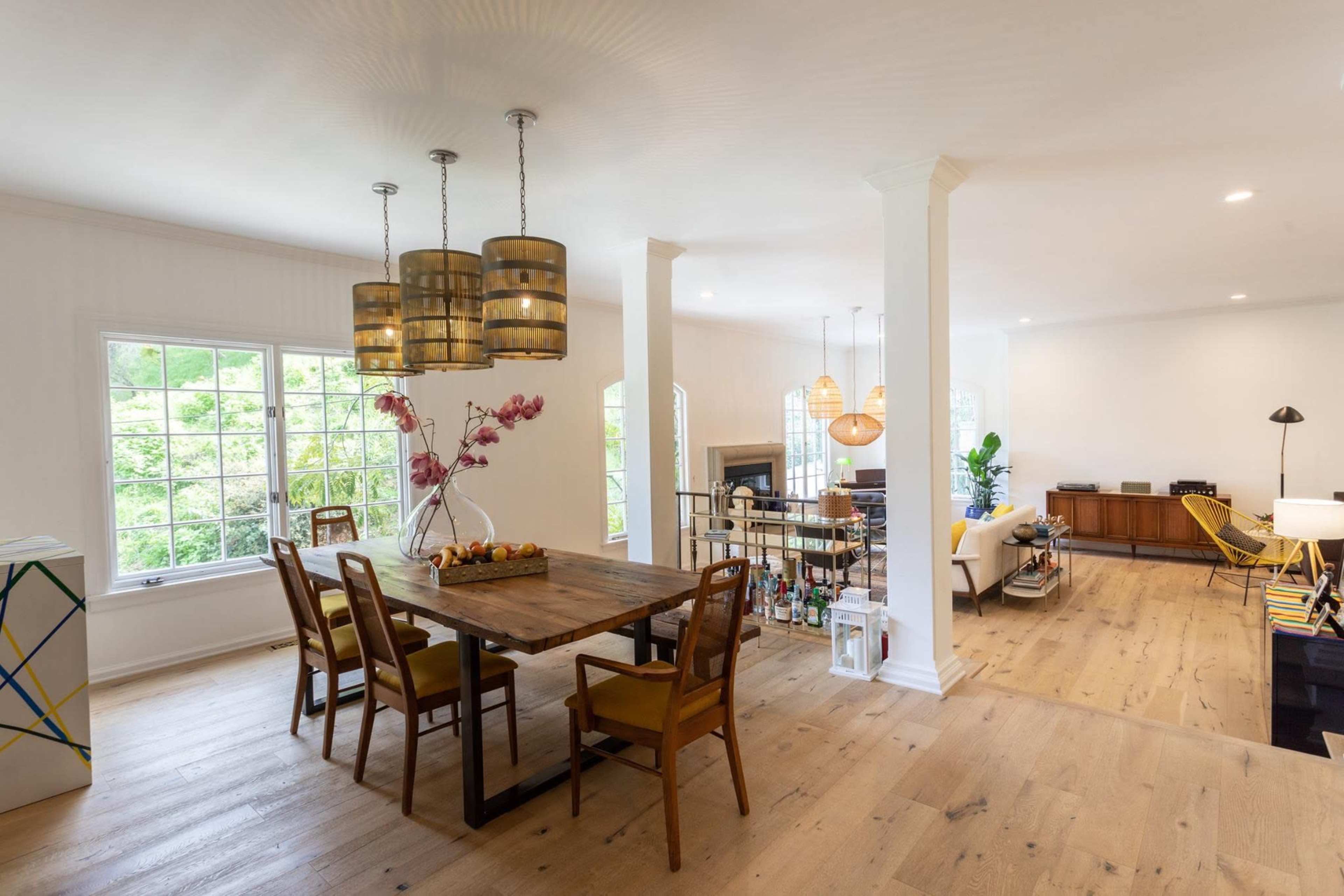 The image shows a spacious dining area with a wooden table surrounded by chairs, illuminated by pendant lights, and featuring a large window with greenery outside.