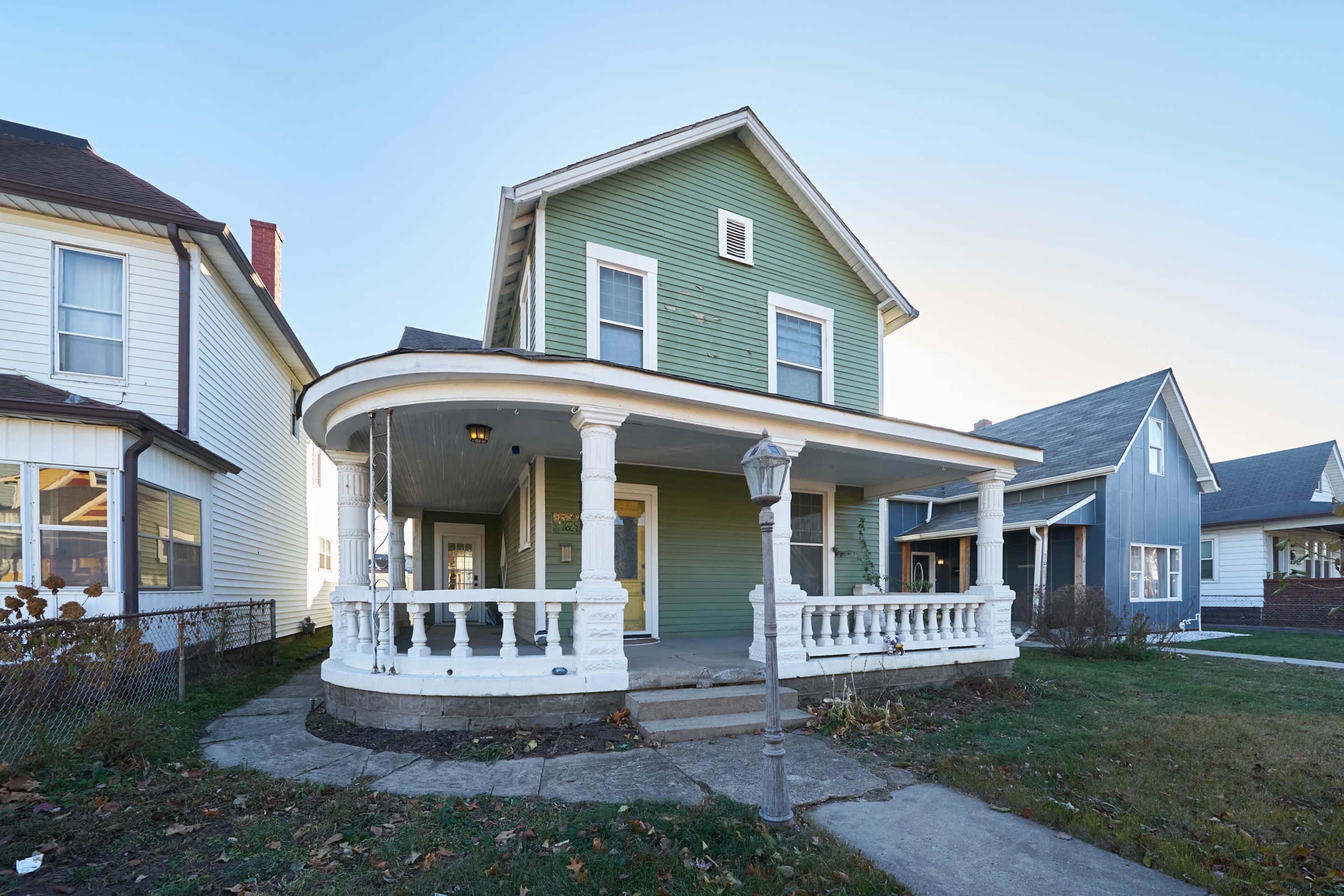 A two-story green house with a front porch featuring white railings and a lamppost on a concrete walkway.