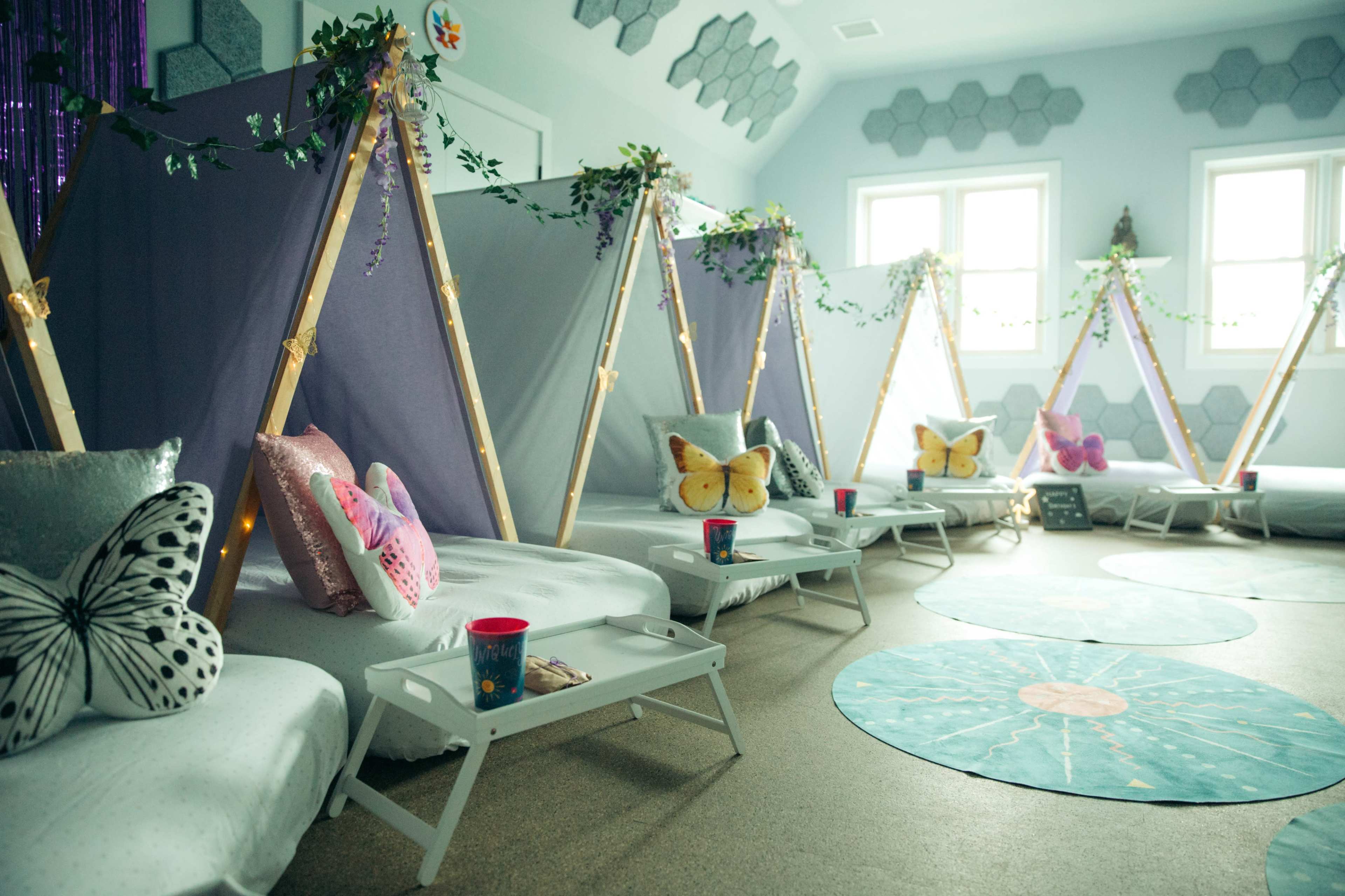A cozy indoor setup featuring teepee-style tents, with white bedding and decorative butterfly pillows, arranged in a brightly lit room.