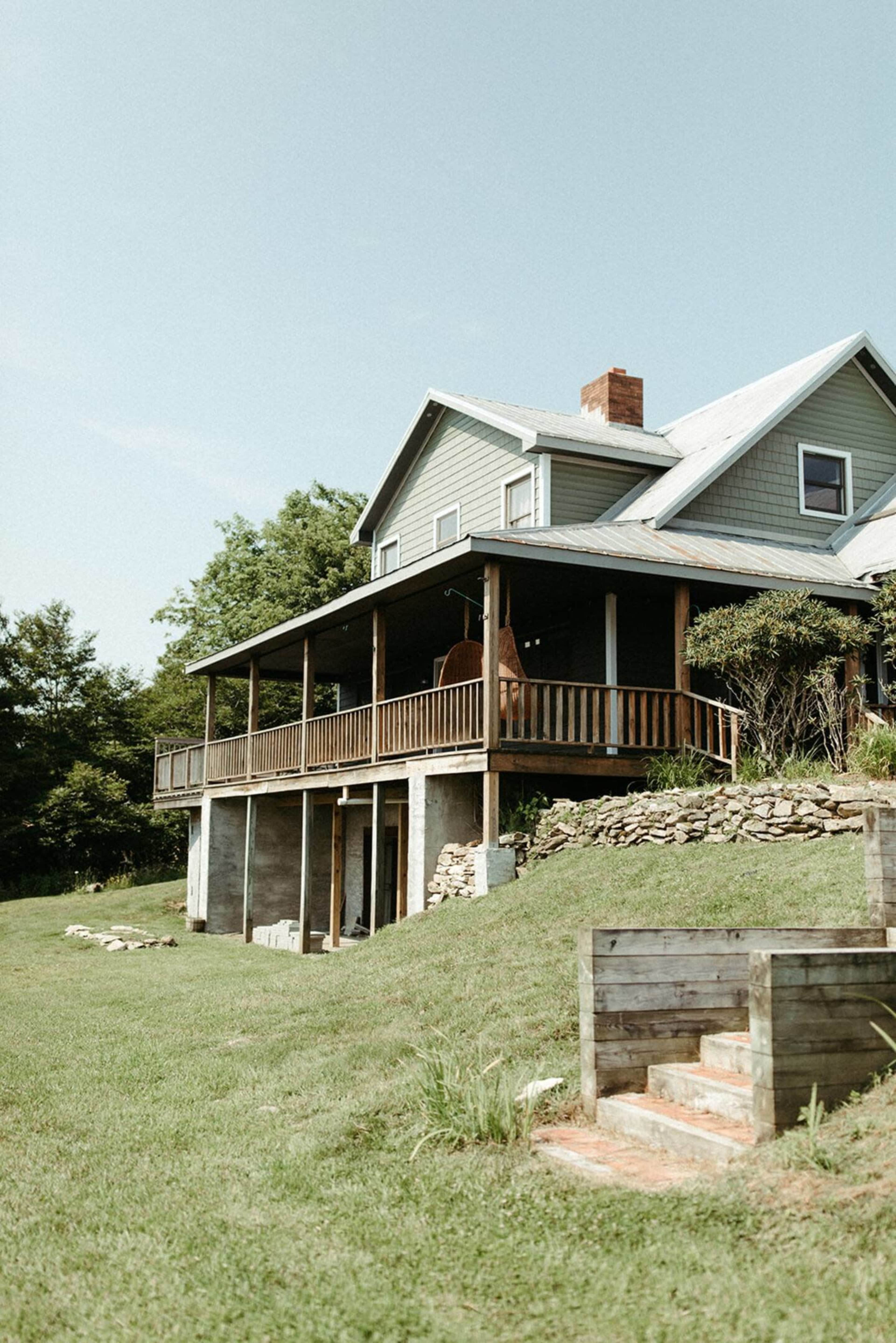 A two-story house with a wooden porch and stone foundation stands on a grassy hillside.