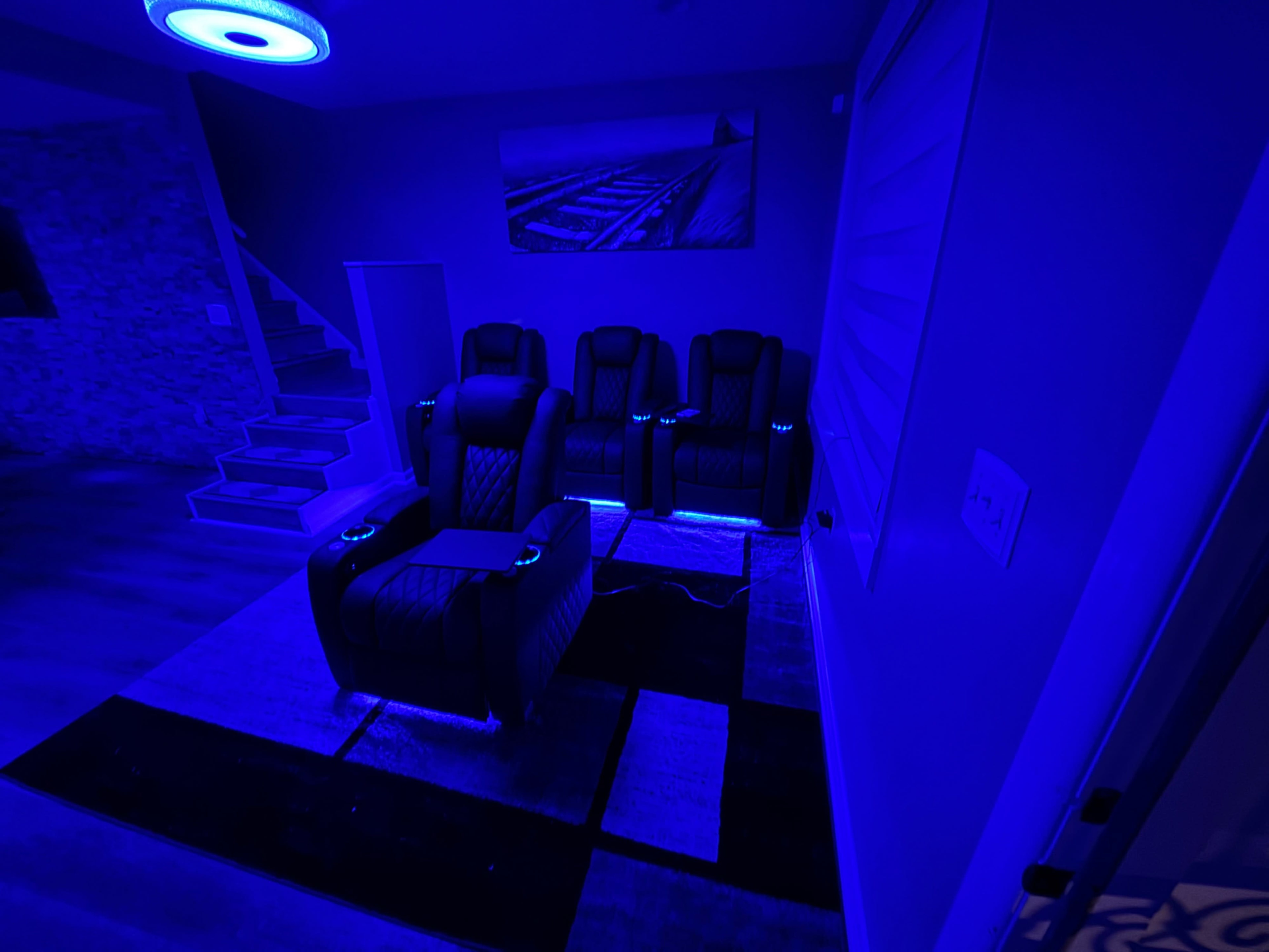 The image shows a dark room with blue lighting, featuring a set of black lounge chairs arranged on a patterned rug, and a staircase visible in the background.