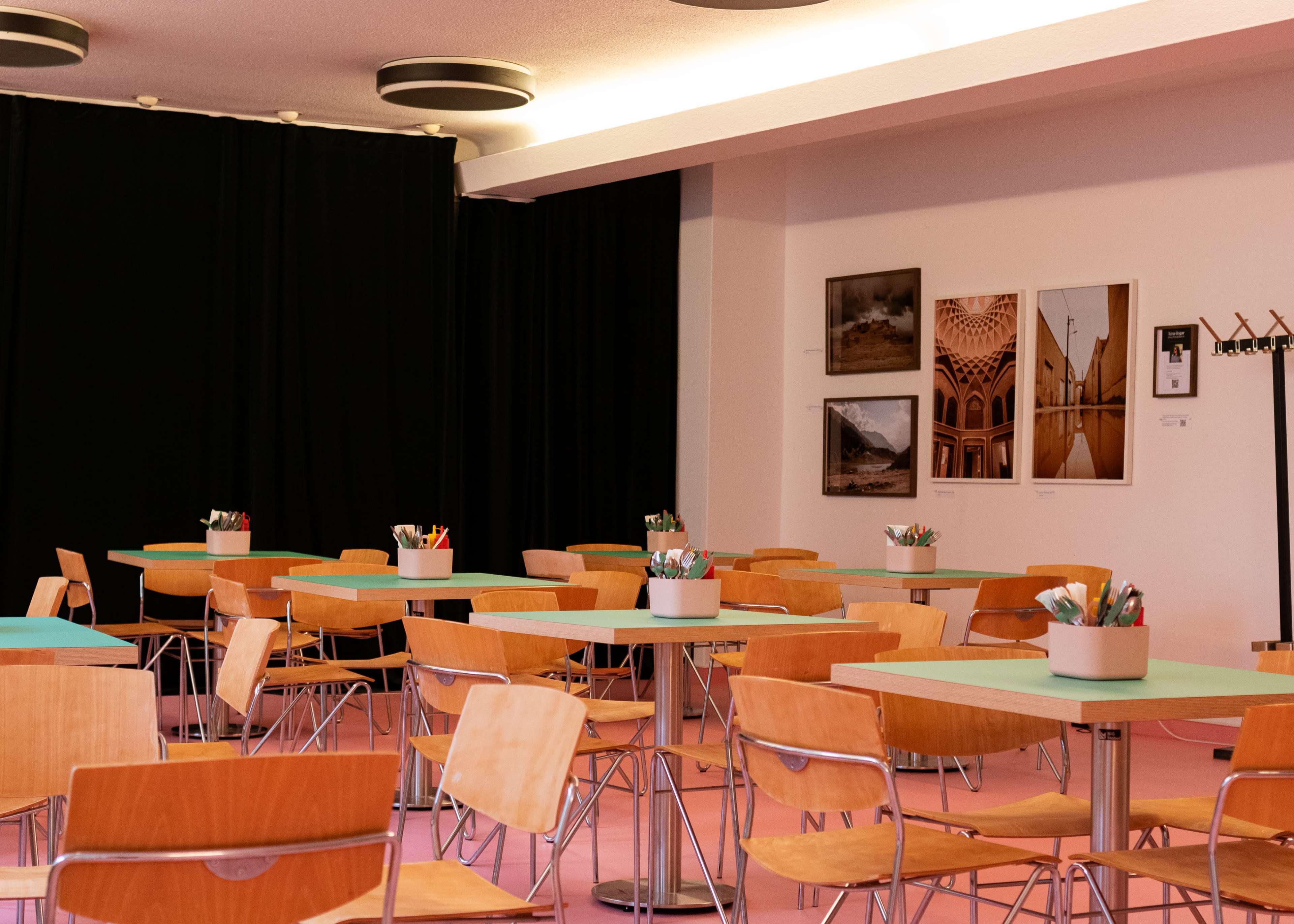 The image shows a brightly lit room with several wooden tables and chairs, featuring potted plants and art on the walls.