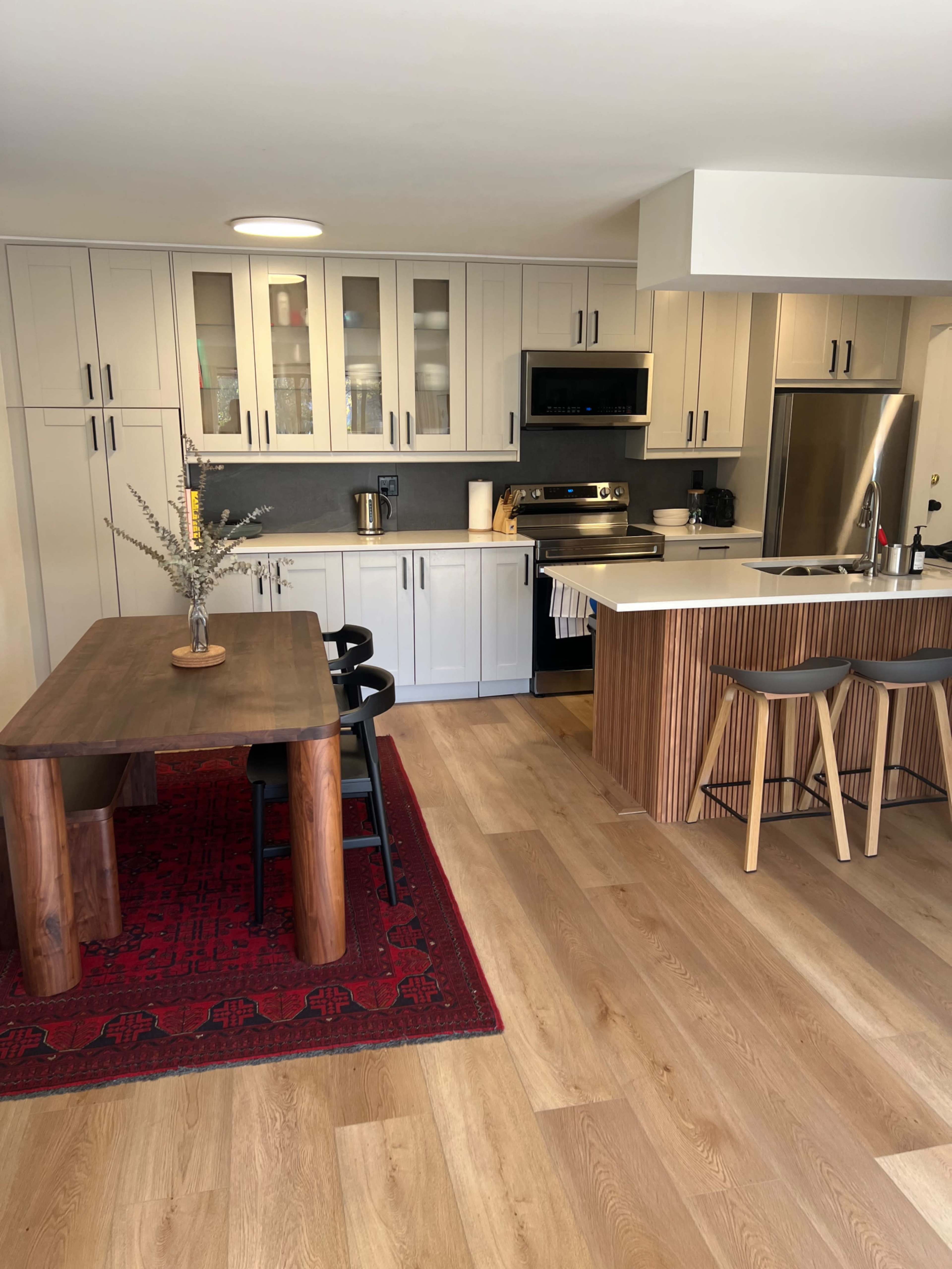 A modern kitchen features wooden cabinetry, stainless steel appliances, a dining table with black chairs, and a red area rug on the floor.