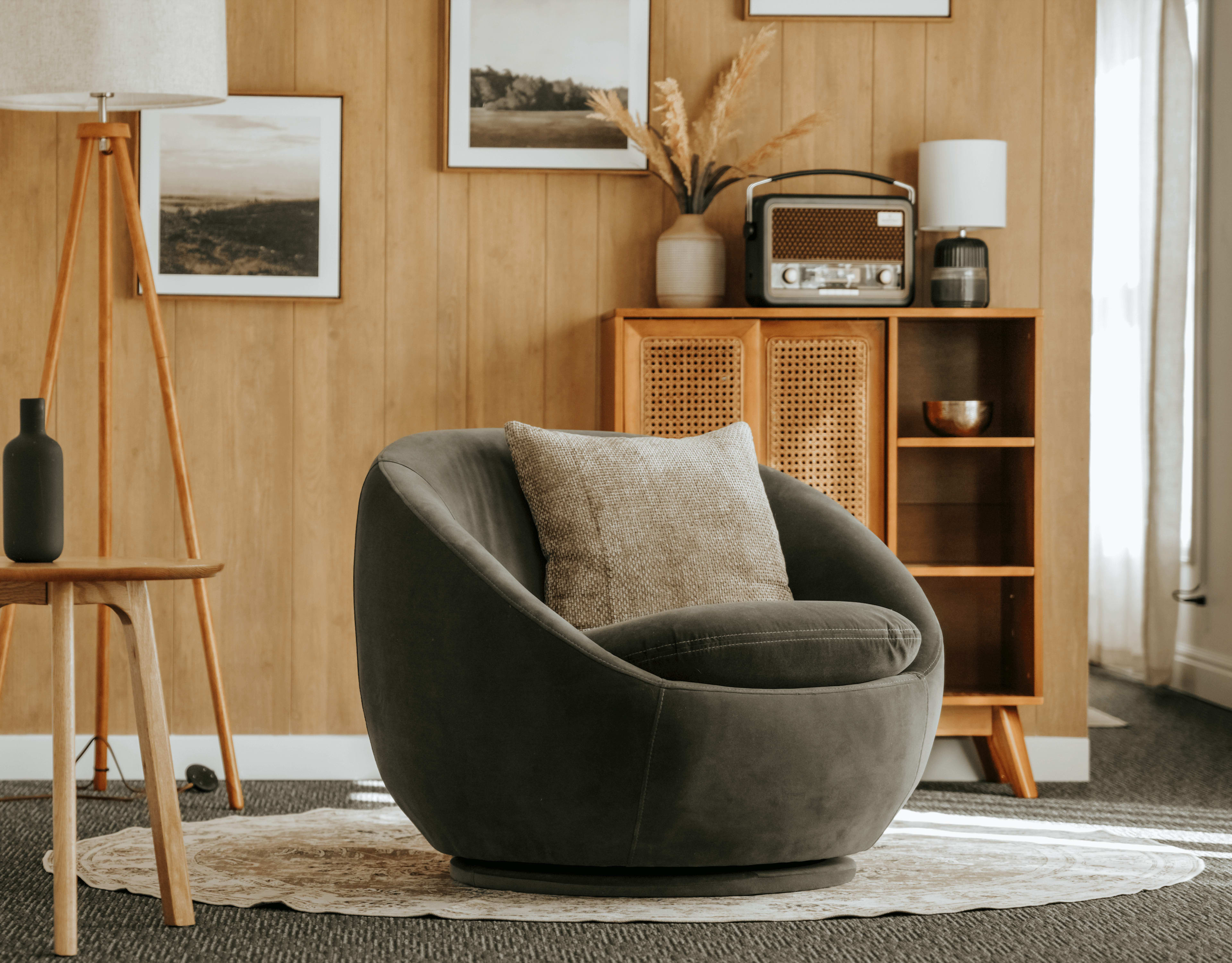 A modern, round chair with a cushion sits on a decorative rug in a room featuring wooden paneling and framed artwork.