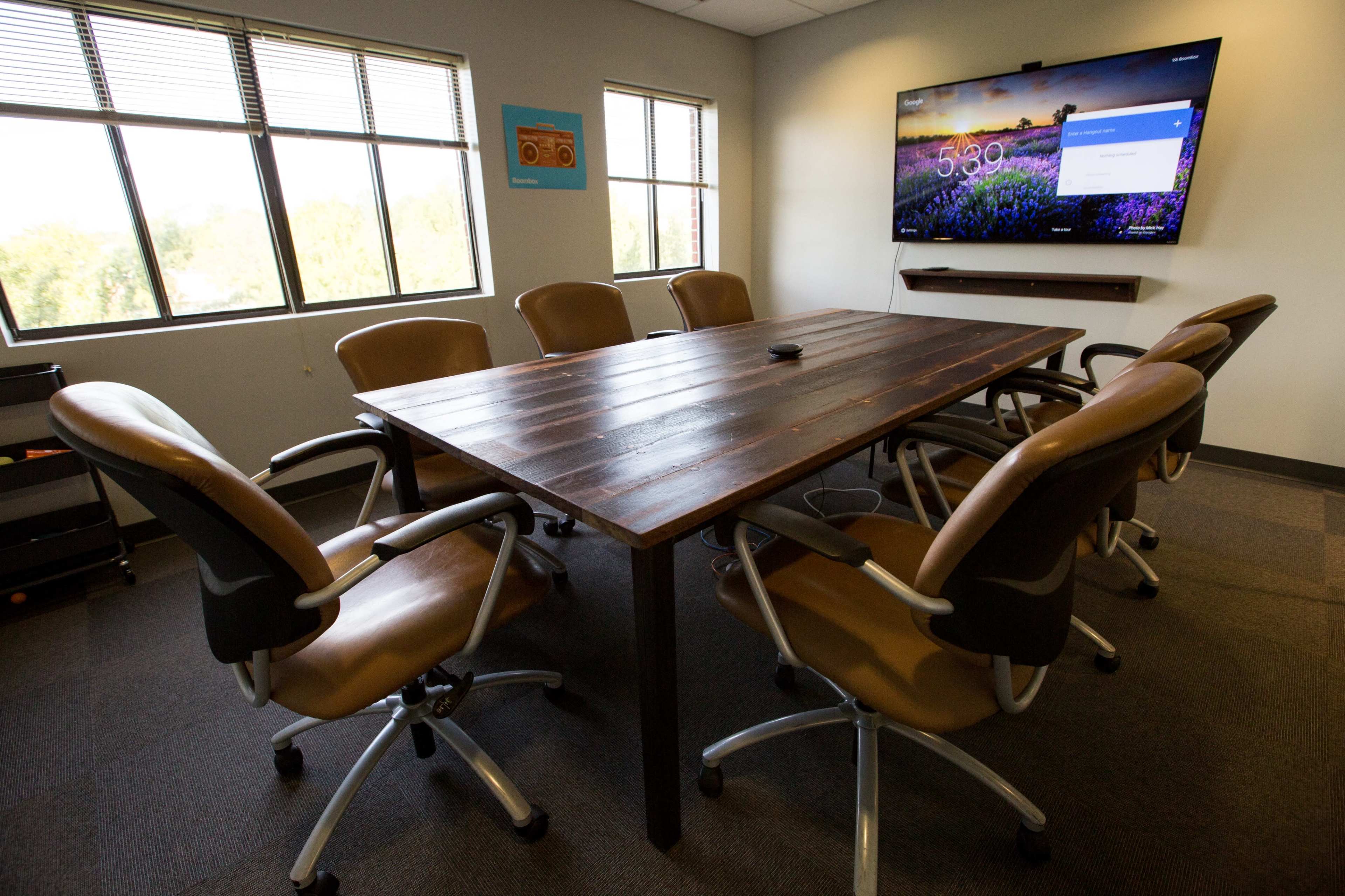 A conference room features a large wooden table surrounded by ergonomic chairs, with a television mounted on the wall displaying the time.