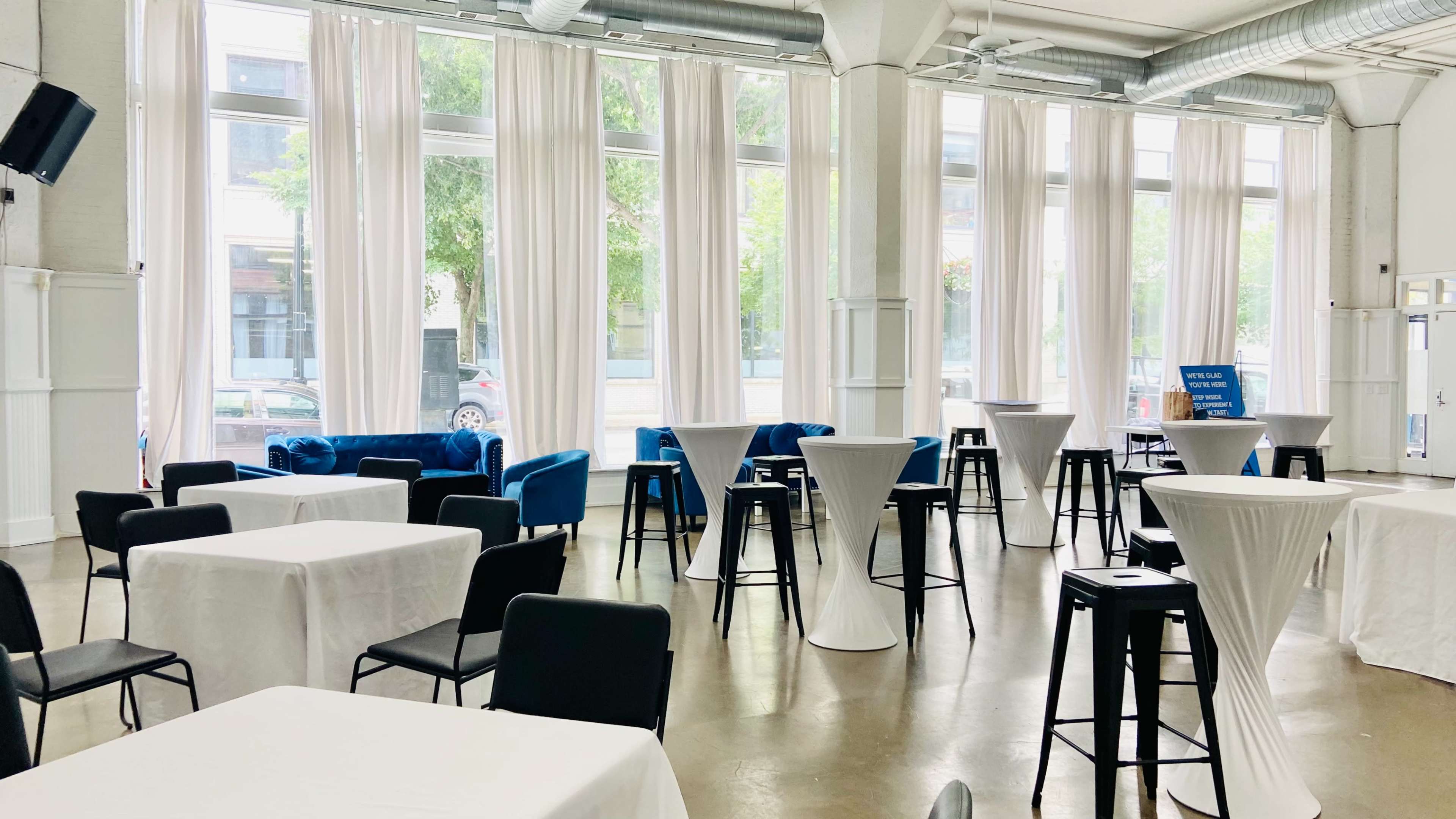 A spacious event venue with tall windows, white drapes, and various tables arranged throughout the room.