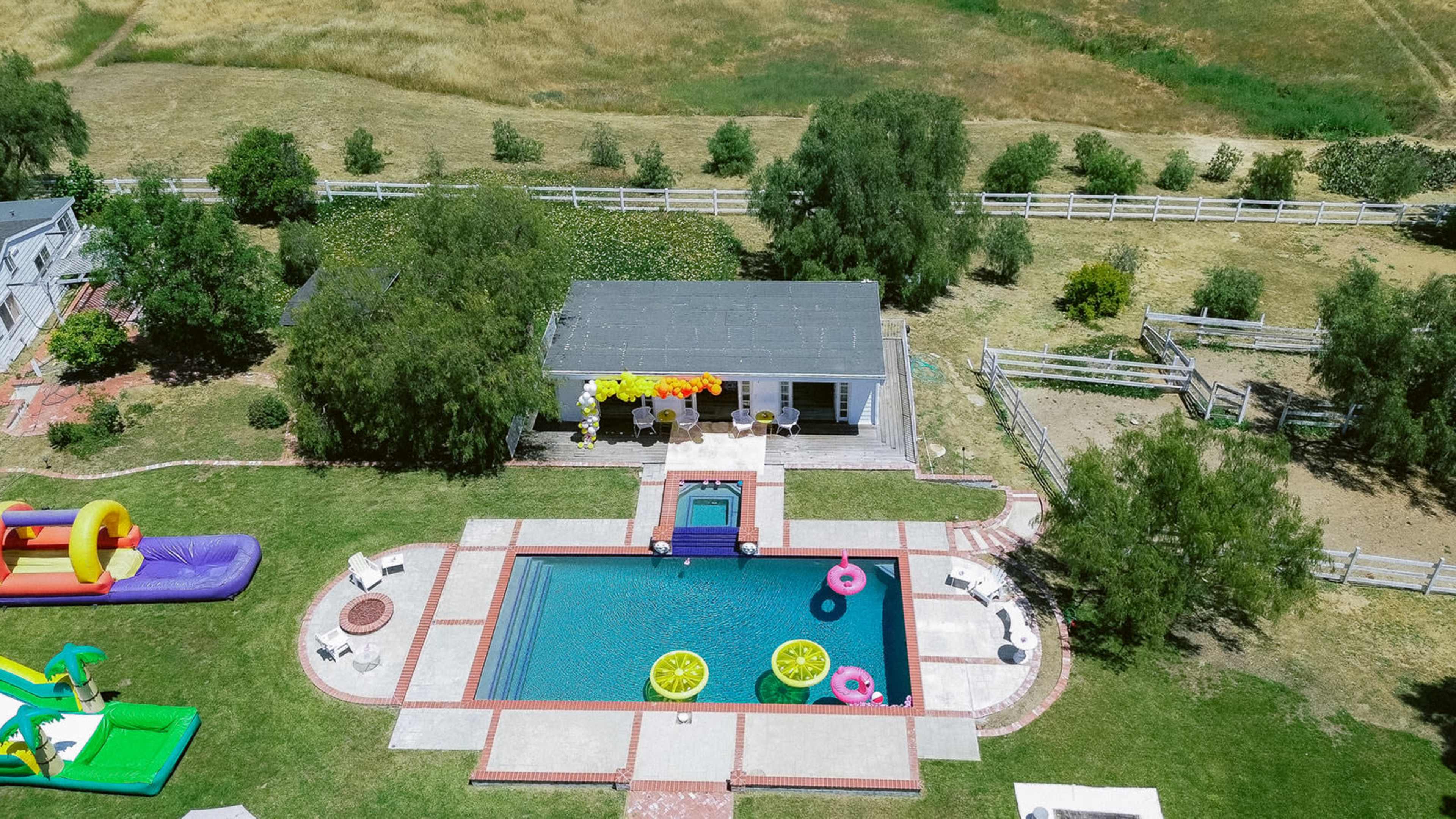 An aerial view showcases a rectangular swimming pool surrounded by colorful pool floats, a small house with a covered patio, and green landscaped areas.