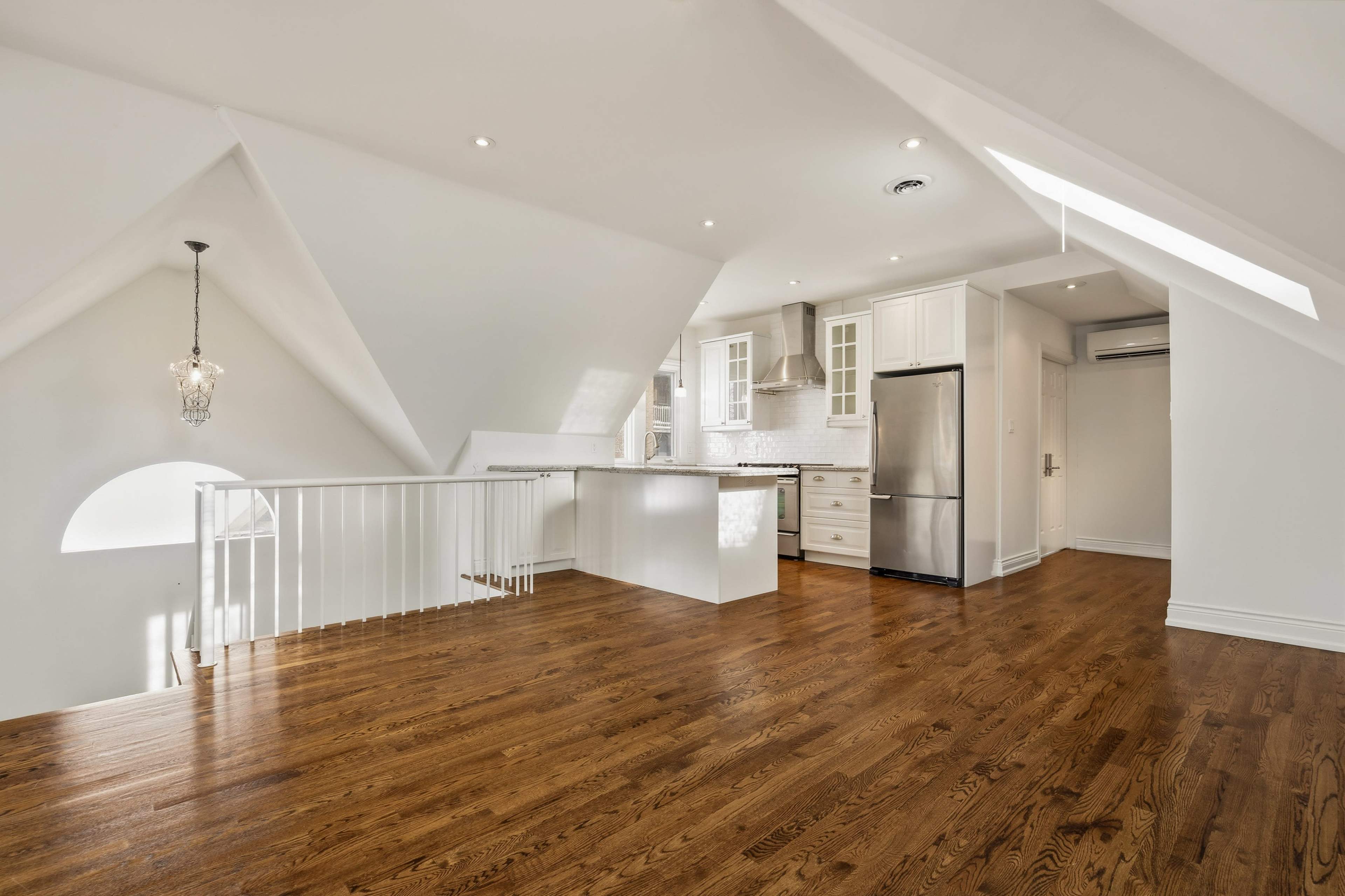 The image shows a well-lit, modern open-concept kitchen and living area with hardwood floors and angled white ceilings.