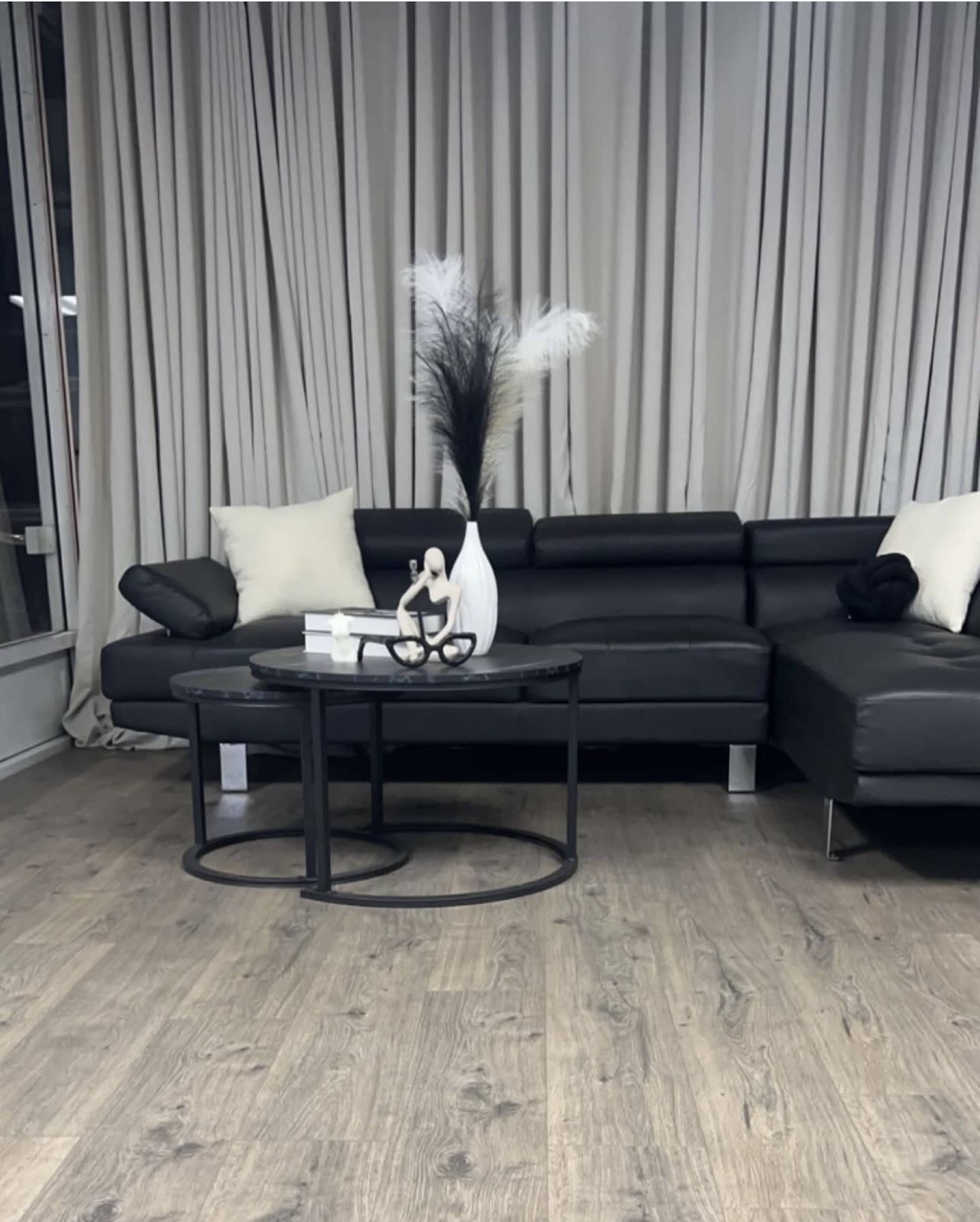 The image shows a modern living room featuring a black leather sectional sofa, a round black coffee table, and decorative elements including a white vase and tall feathers against a backdrop of light curtains.