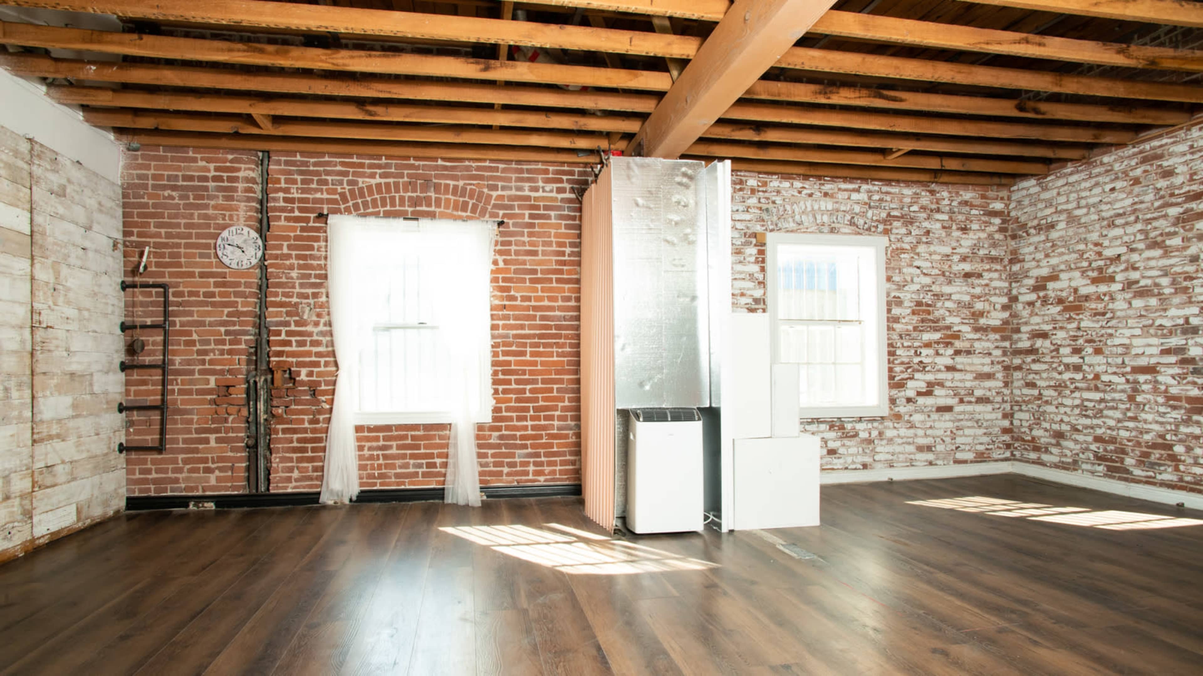 A spacious room with exposed brick walls, wooden beams overhead, and a modern fridge positioned near a window.