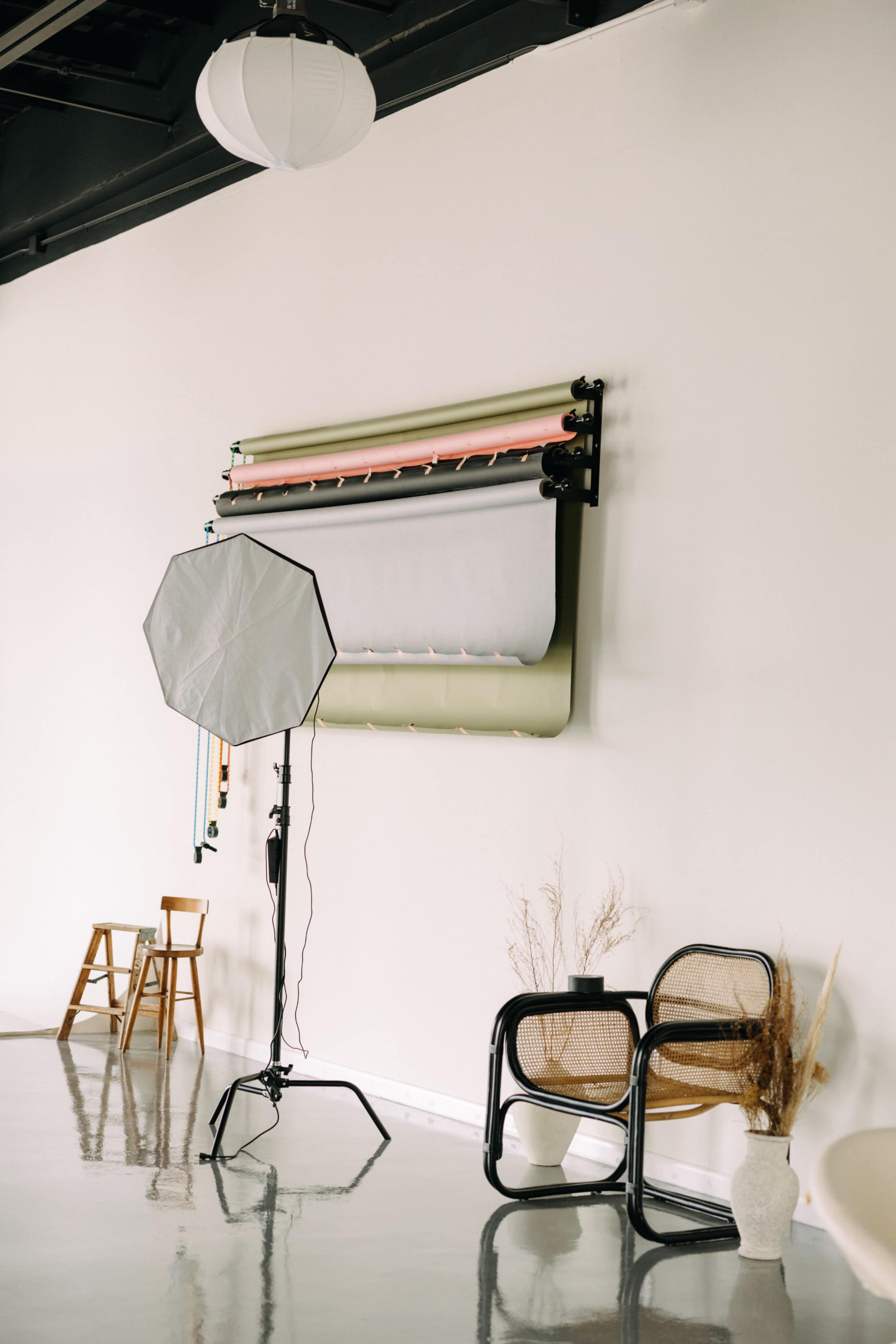 A minimalist studio setup with a backdrop roller, a photography light stand, and two different styles of chairs against a plain wall.