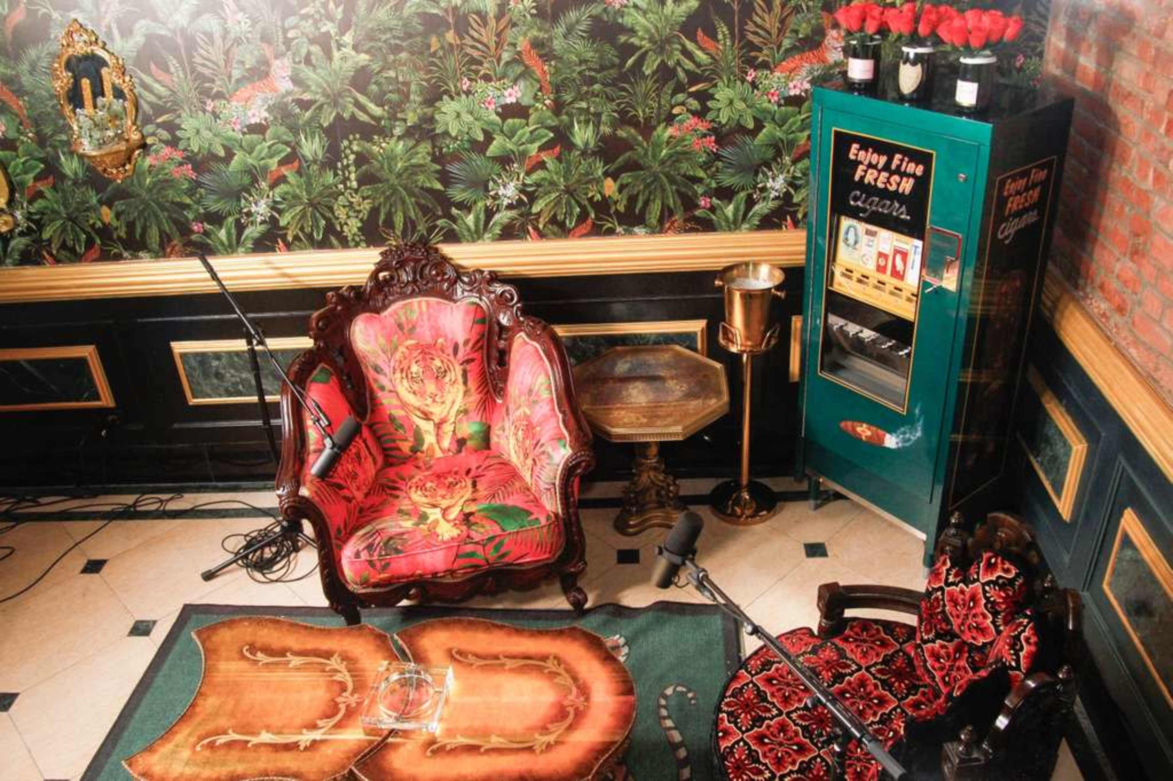 The scene features an elegantly decorated room with two vintage armchairs, a small side table, a cigarette vending machine, and lush floral wallpaper.